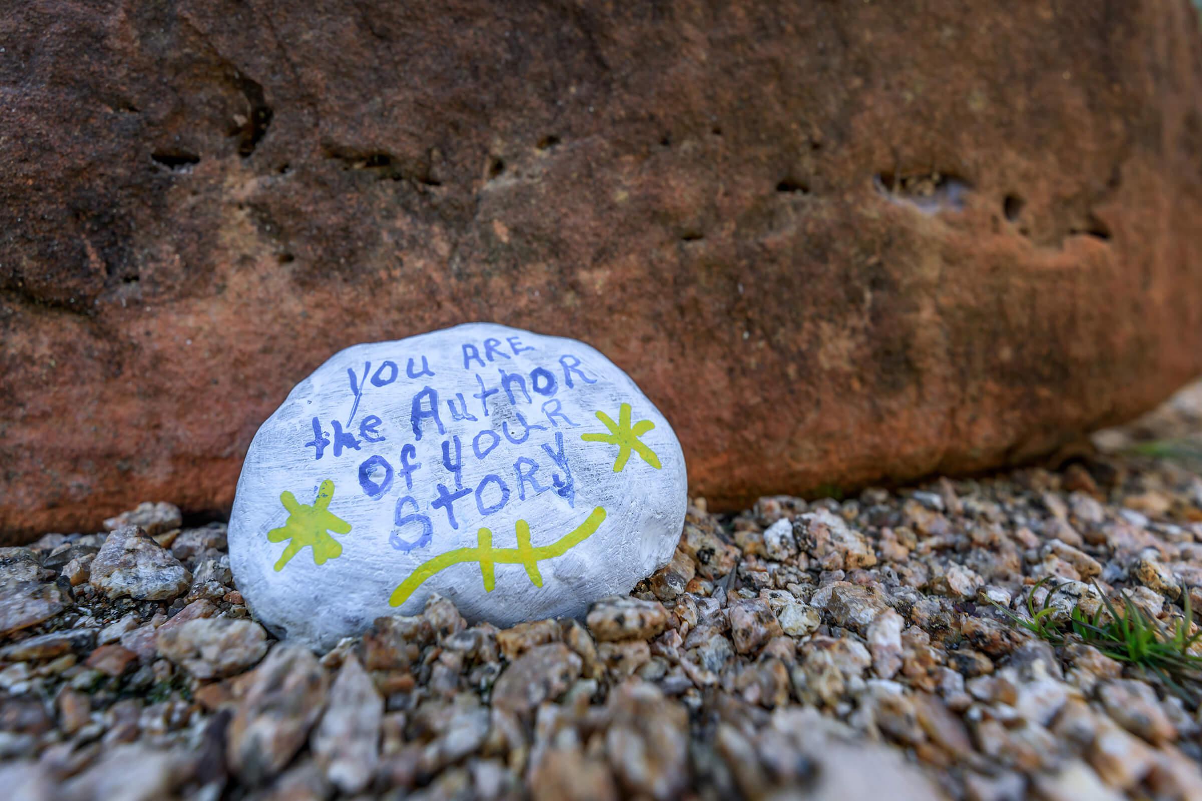 A painted rock resting on gravel features the inspirational quote "You are the author of your story" in black ink, surrounded by green star-like designs. The rock is positioned near a larger stone, suggesting a natural outdoor setting.