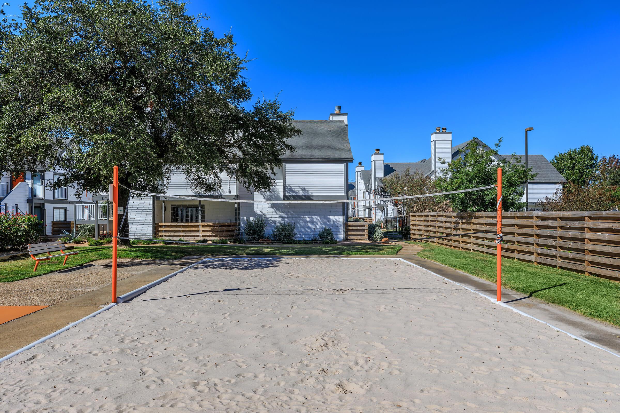 A sandy volleyball court with orange posts, surrounded by green grass and wooden fencing. In the background, there are two white residential buildings and a blue sky. A bench is visible to the left of the court, creating a peaceful recreational area.