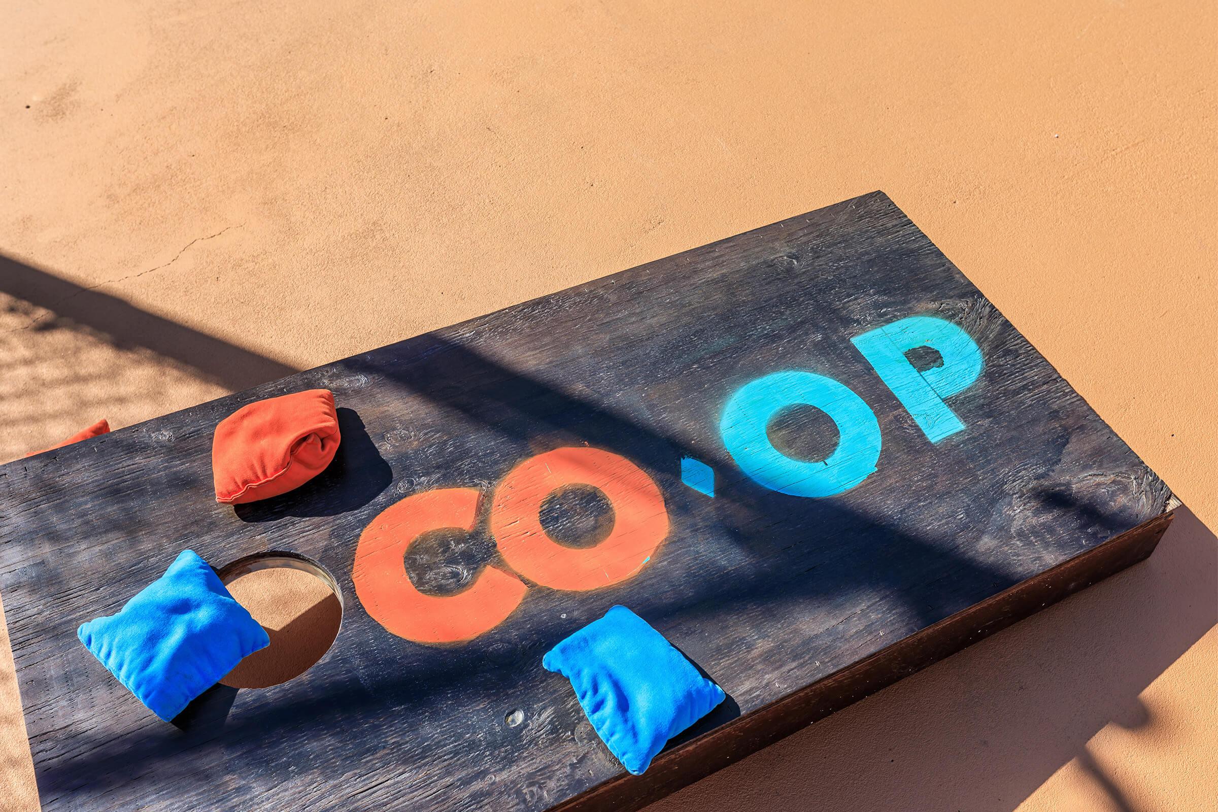 A wooden cornhole board with the word "CO-OP" painted in orange and blue. Two colorful bean bags, one red and one blue, are resting on the board. The background features a sandy surface with shadows cast over the board, suggesting a sunny outdoor setting.