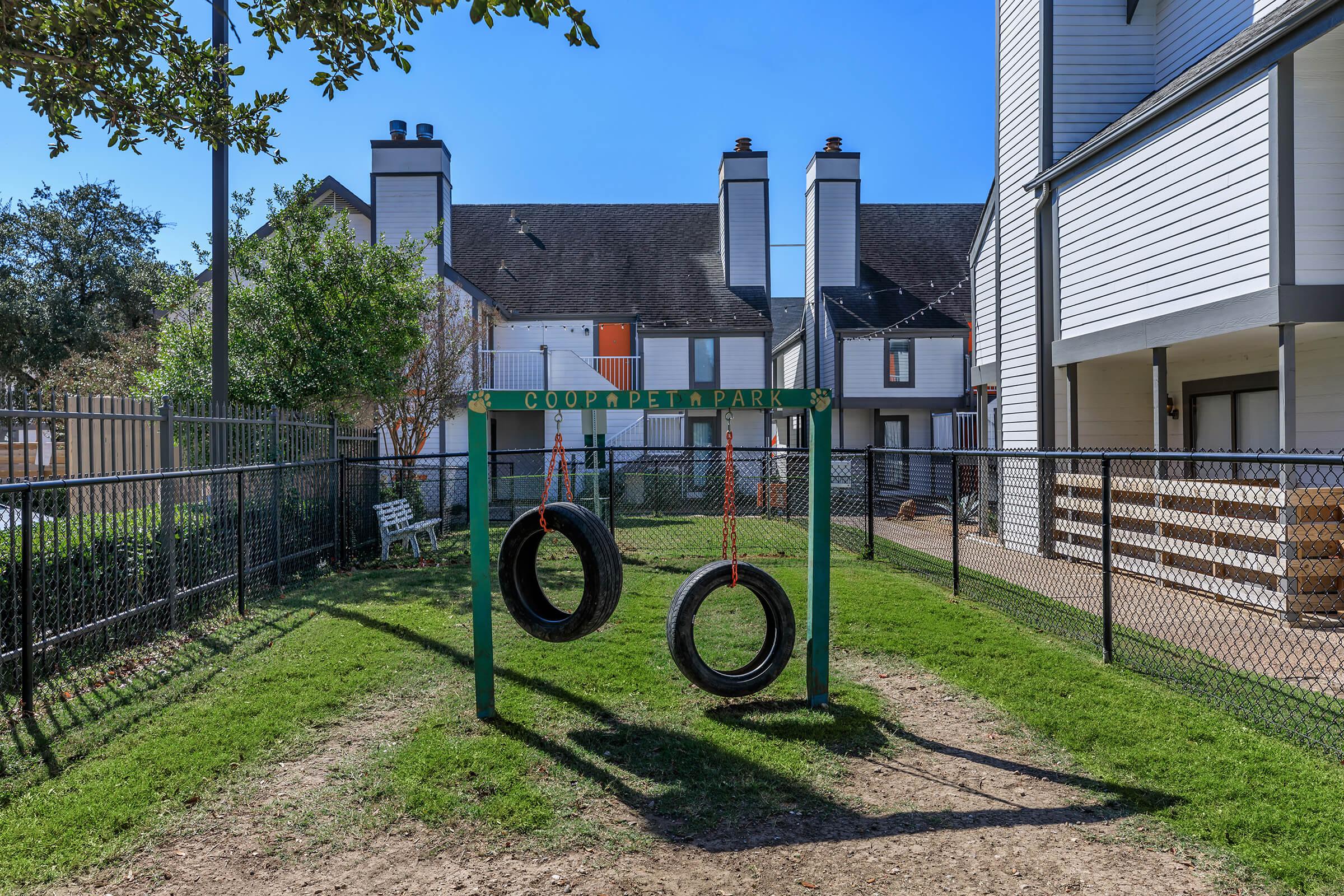 A swing set made of old tires, suspended by ropes, is situated in a grassy area within a fenced park. Surrounding the park are residential buildings with a clear blue sky above. Trees provide some greenery, and a bench is visible in the background.