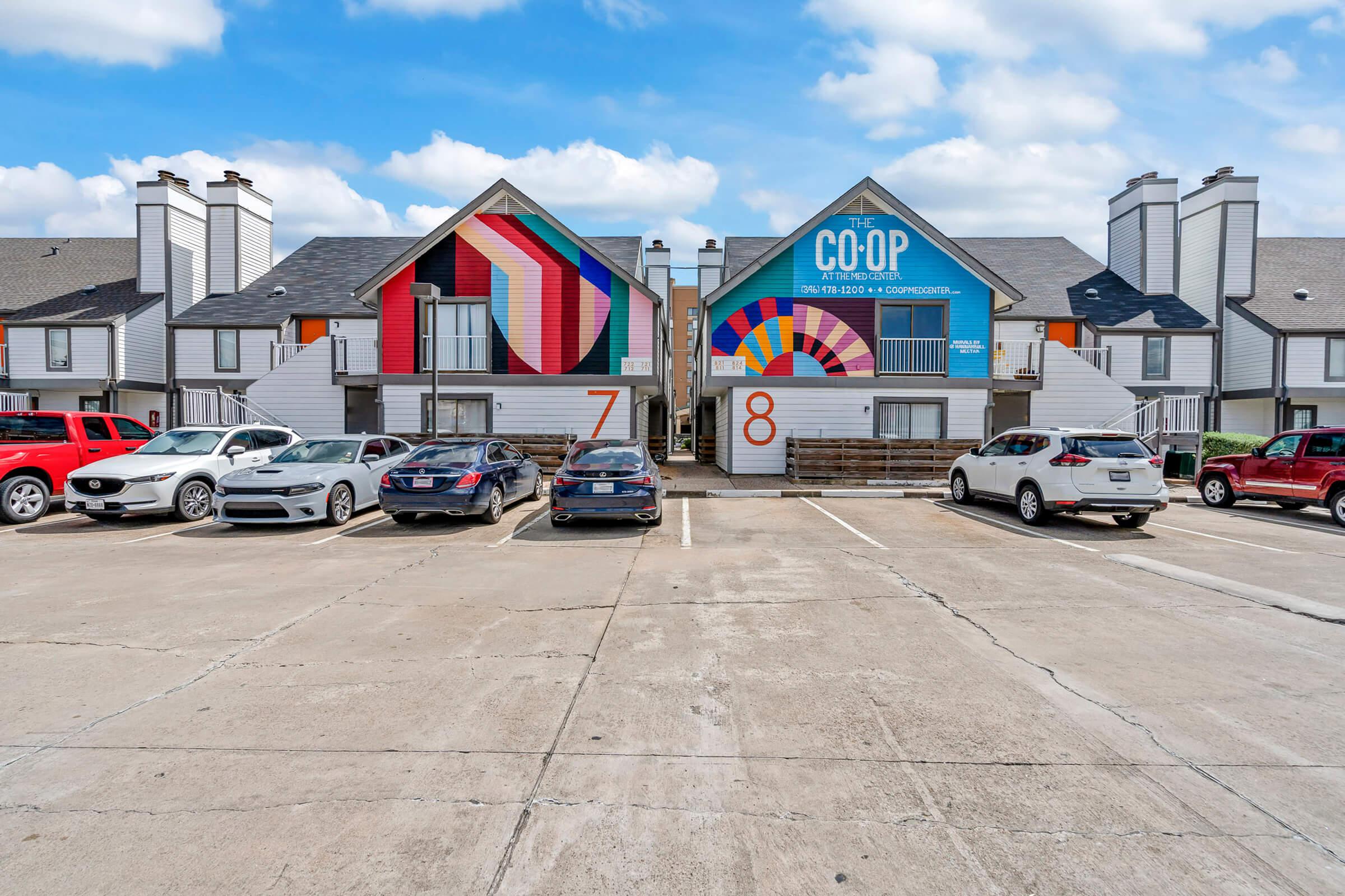 A parking lot in front of two apartment buildings, numbered 7 and 8. The building on the right features a colorful mural that reads "The Co-op." Several cars are parked in the lot, and the sky is partly cloudy, giving a bright ambiance to the scene.