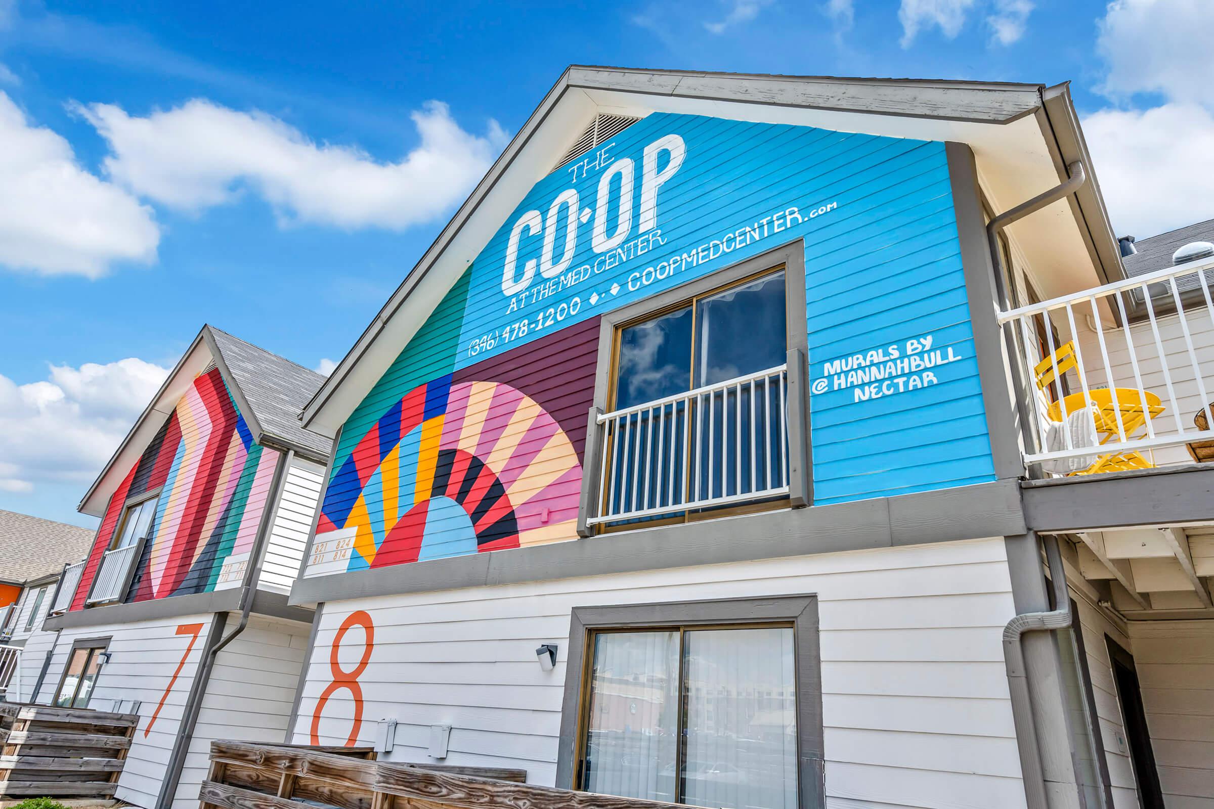 Colorful exterior of a building identified as "The Co-op" with murals and a bright design. The address and operation hours are displayed prominently, along with the names of the mural artists. The building features a mix of modern architecture with decorative elements, surrounded by a clear blue sky.