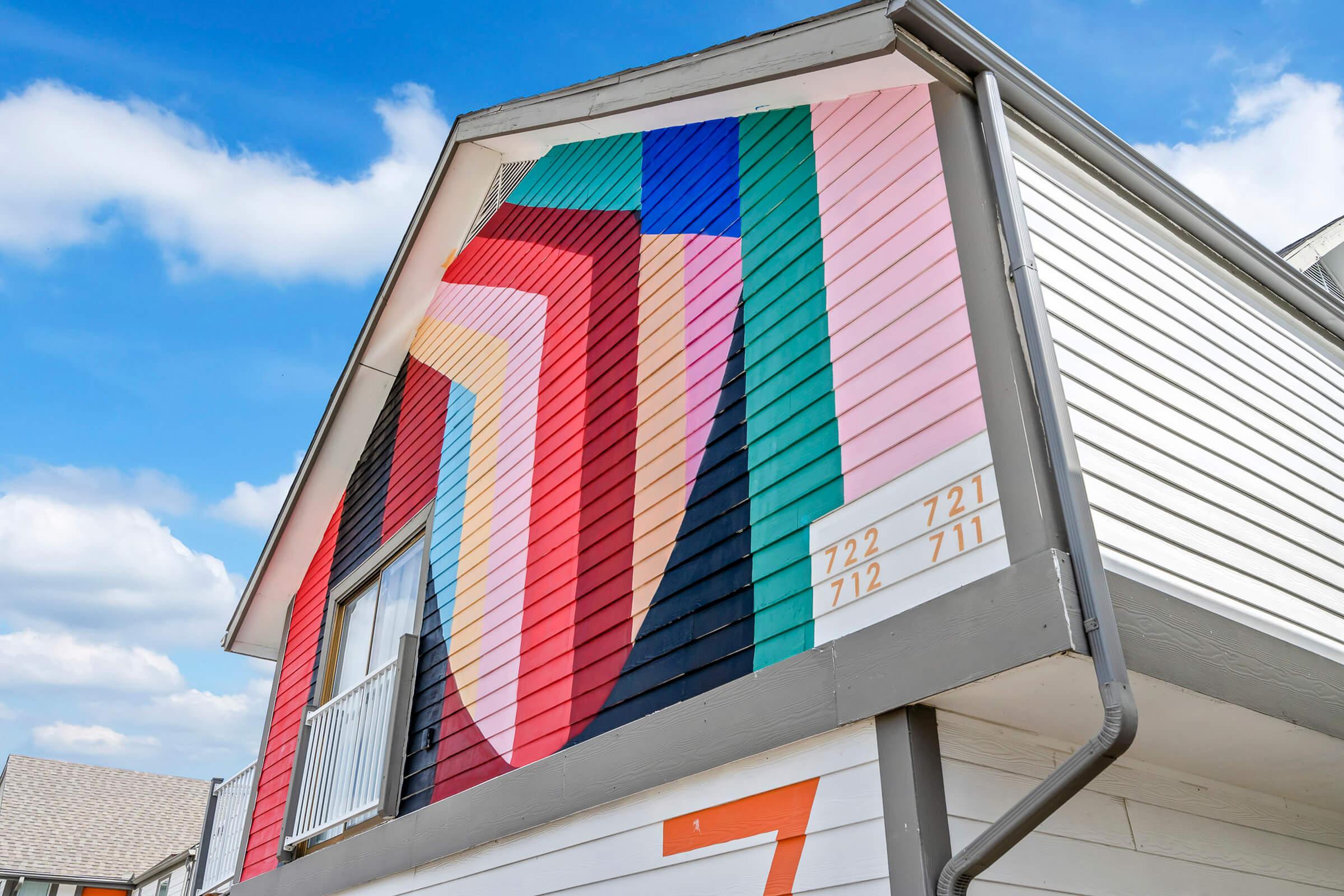 A vibrant mural on the side of a building, featuring geometric patterns in shades of red, blue, pink, and green. The design creates a dynamic visual impact against a backdrop of a cloudy sky. The building also has house numbers displayed on the side.