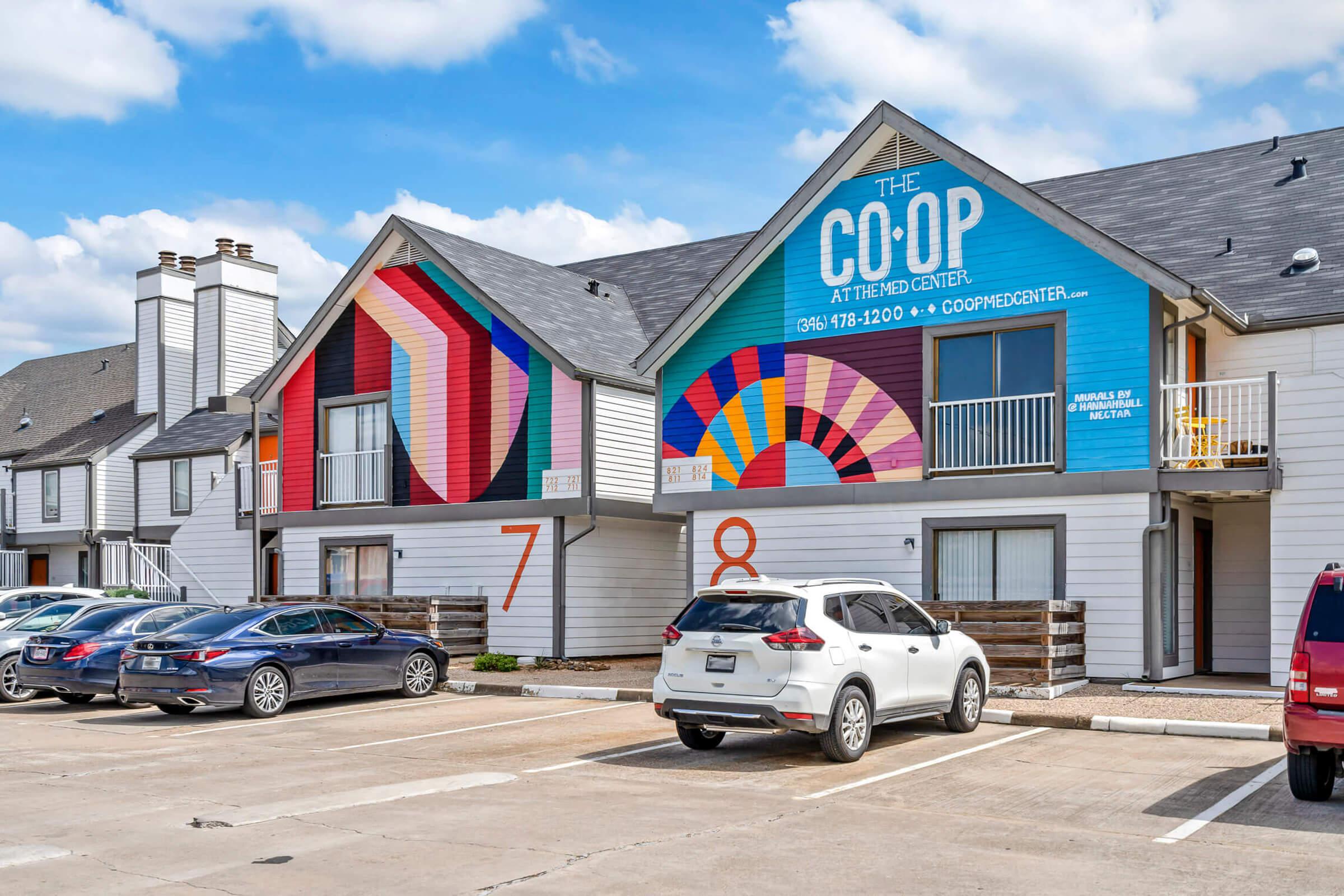 A colorful apartment community featuring modern murals on the exterior. The murals display geometric shapes in vibrant colors. Two units are visible, numbered 7 and 8, with parked cars in the foreground. The buildings have a sleek, contemporary design with gray roofing. A bright blue sign indicates the location as “The Co-Op.”