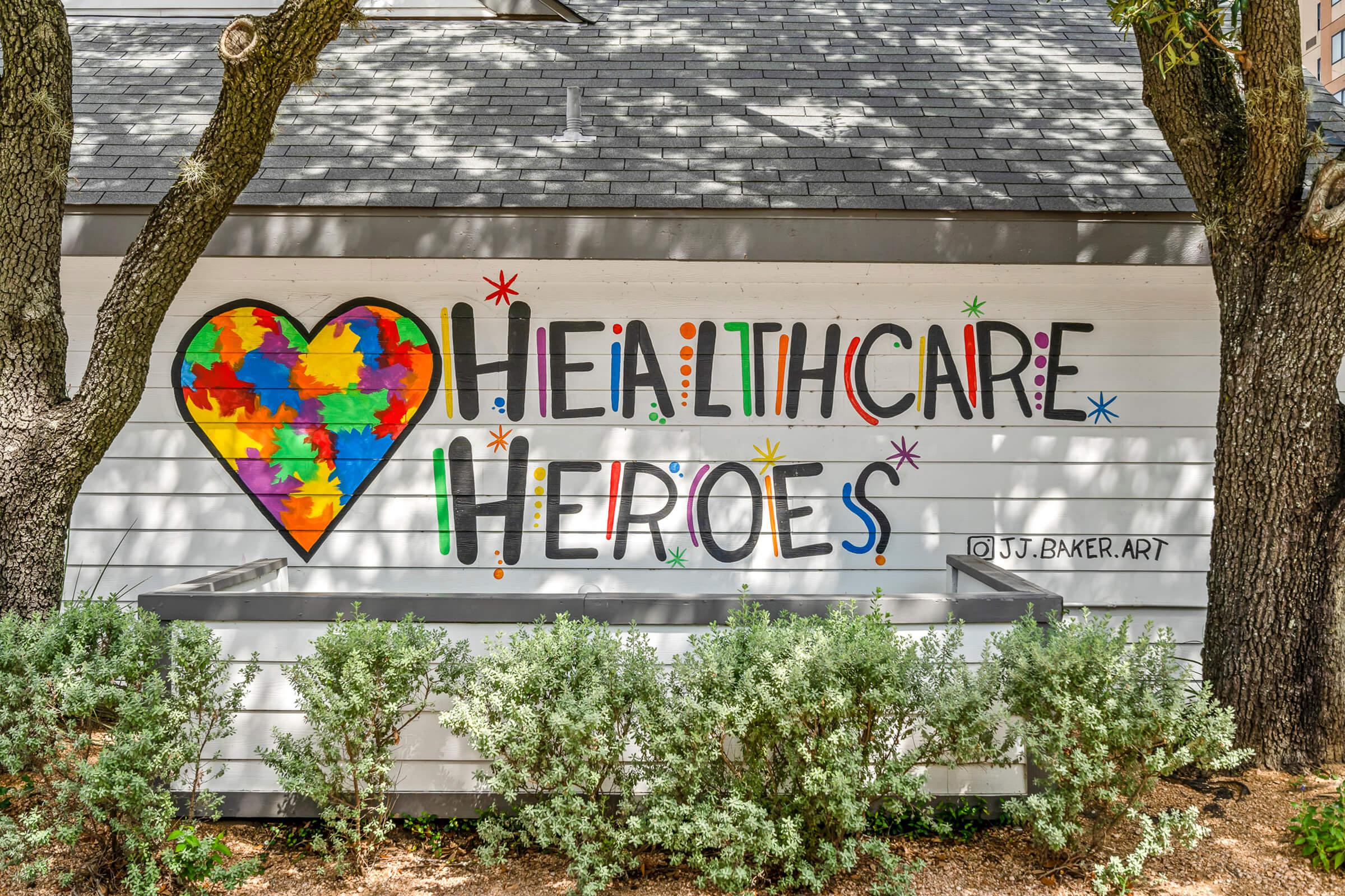 A colorful mural on a white wall featuring a heart made of puzzle pieces, accompanied by the text "Healthcare Heroes" in bold letters. The artwork includes bright stars and playful designs, highlighting appreciation for healthcare workers. Surrounding greenery adds to the vibrant scene.