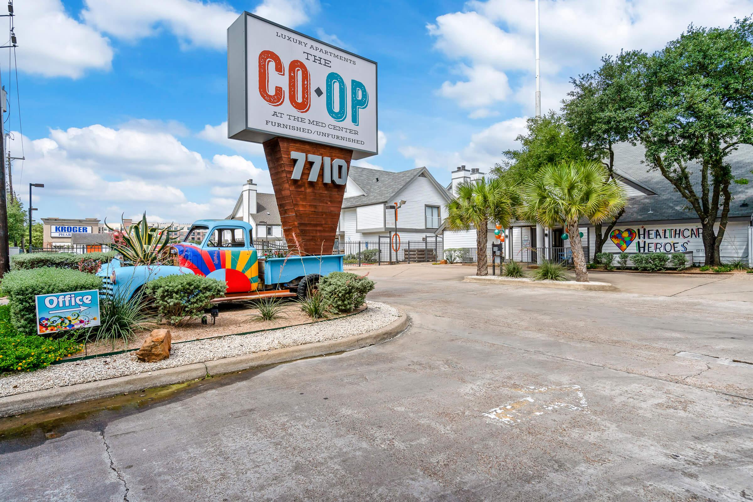 A colorful sign for "CO-OP" at 7710, along with a retro truck painted in bright colors. The entrance features palm trees and various signs advertising office space and a tribute to healthcare heroes, set against a backdrop of blue skies and fluffy clouds.