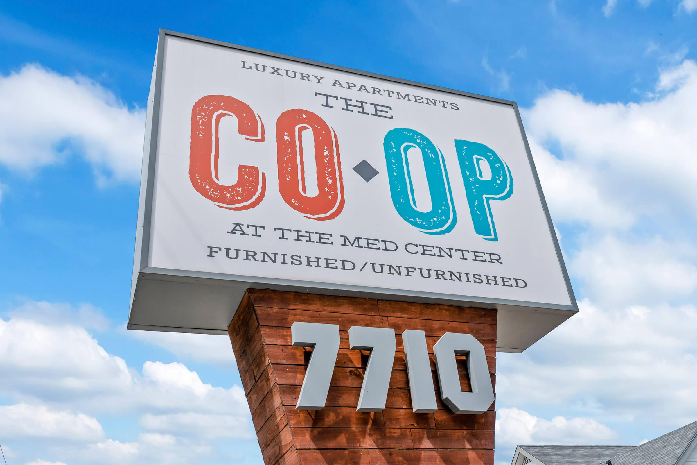 Sign for "The Co-Op" luxury apartments located at the Med Center. It indicates options for furnished or unfurnished units. The sign features a modern design with a wooden base and prominently displays the address "7710." The background includes a partly cloudy blue sky.