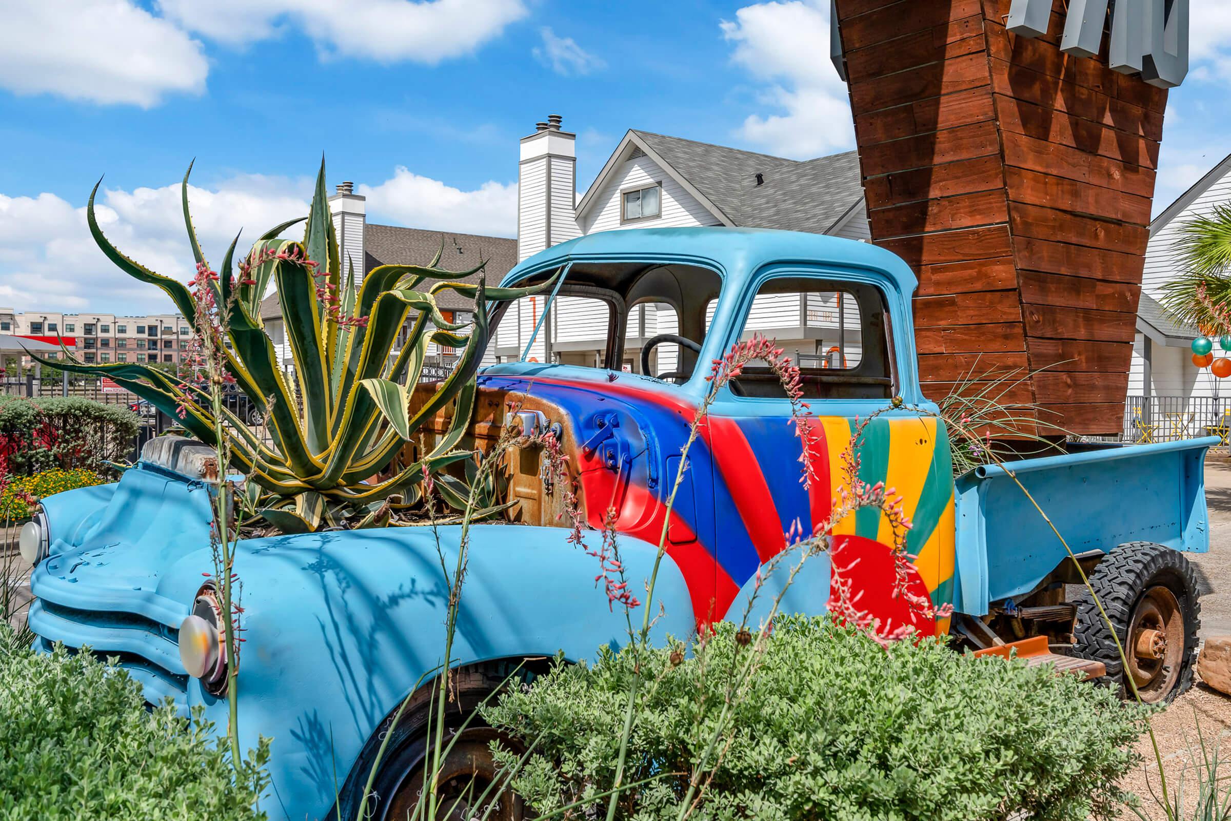 A vintage blue pickup truck with colorful patterns is partially covered by vibrant plants and flowers. It’s situated in a landscaped area with buildings in the background and a bright sky, creating a lively and artistic outdoor setting.