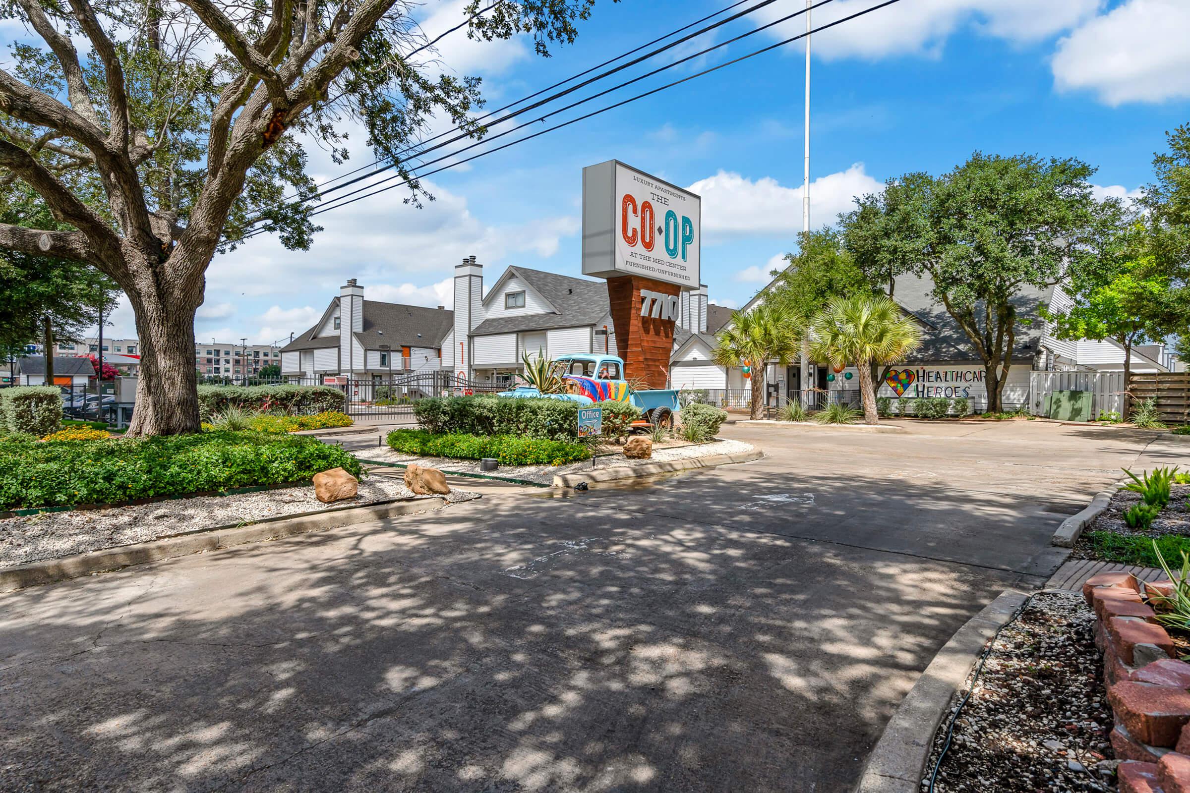 A sunny view of a landscaped entrance to a building complex featuring a large sign that reads "CO-OP." The area includes trees, shrubs, and rocks, with clear blue skies and a few clouds overhead. The scene captures a welcoming atmosphere with well-maintained surroundings.