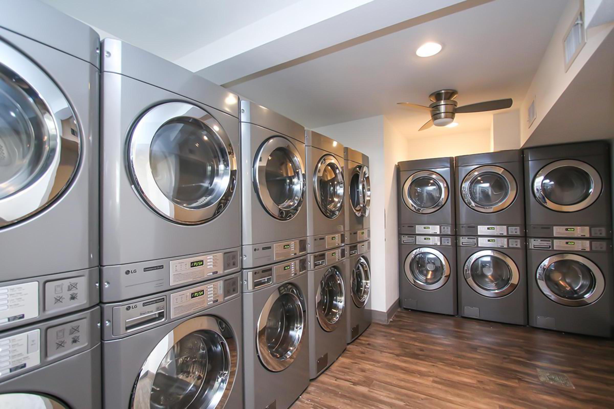 A modern laundry room featuring multiple stacked washer and dryer units in a sleek gray finish. The machines are arranged in two rows against the walls, with a ceiling fan above and stylish wooden flooring, creating a clean and efficient laundry space.