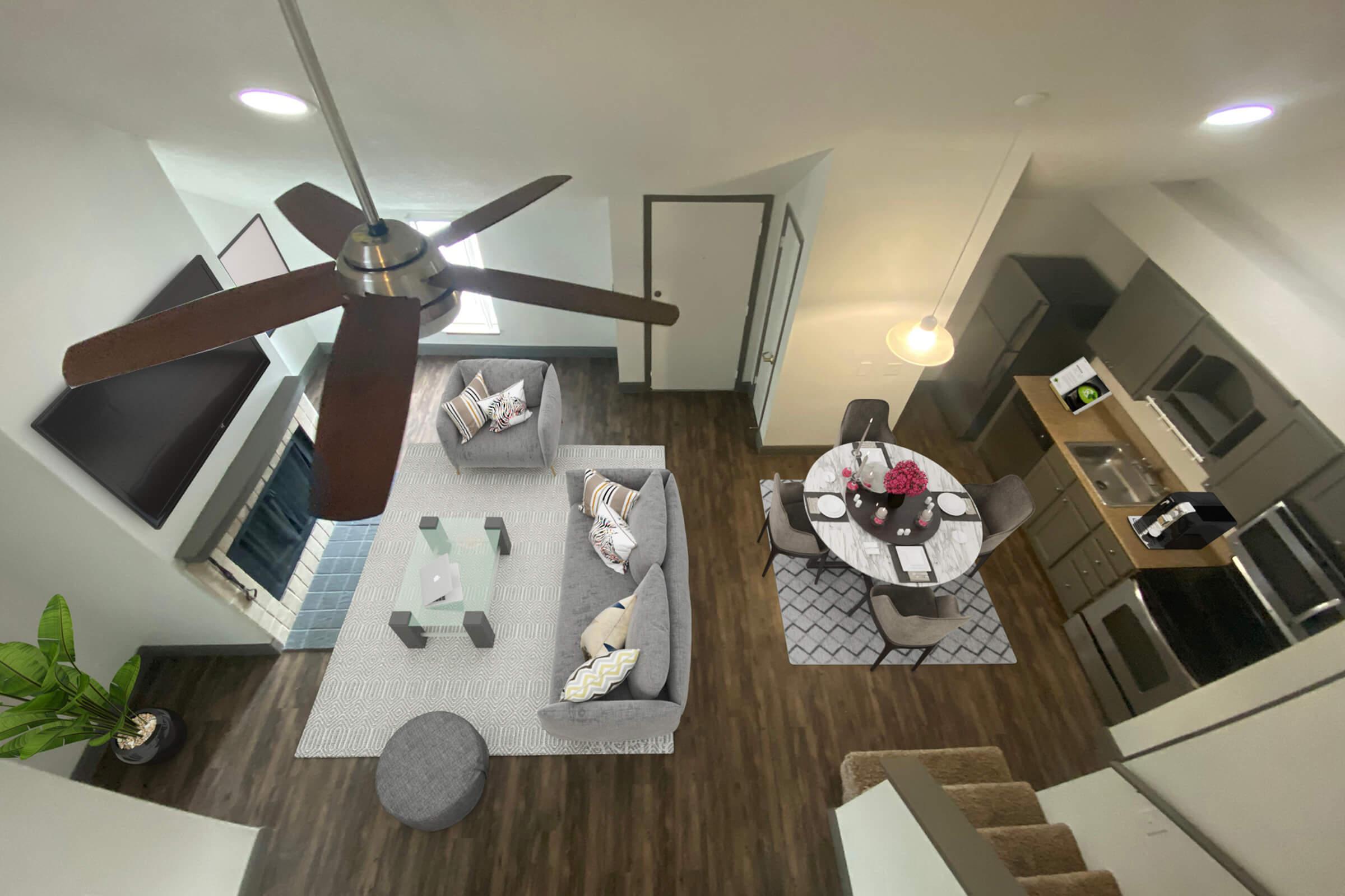 Aerial view of a modern living space featuring a cozy seating area with a gray sofa and decorative pillows, a round dining table with flowers, and a kitchenette. There’s a ceiling fan, a flat-screen TV on the wall, and hardwood flooring. Natural light streams in through large windows.