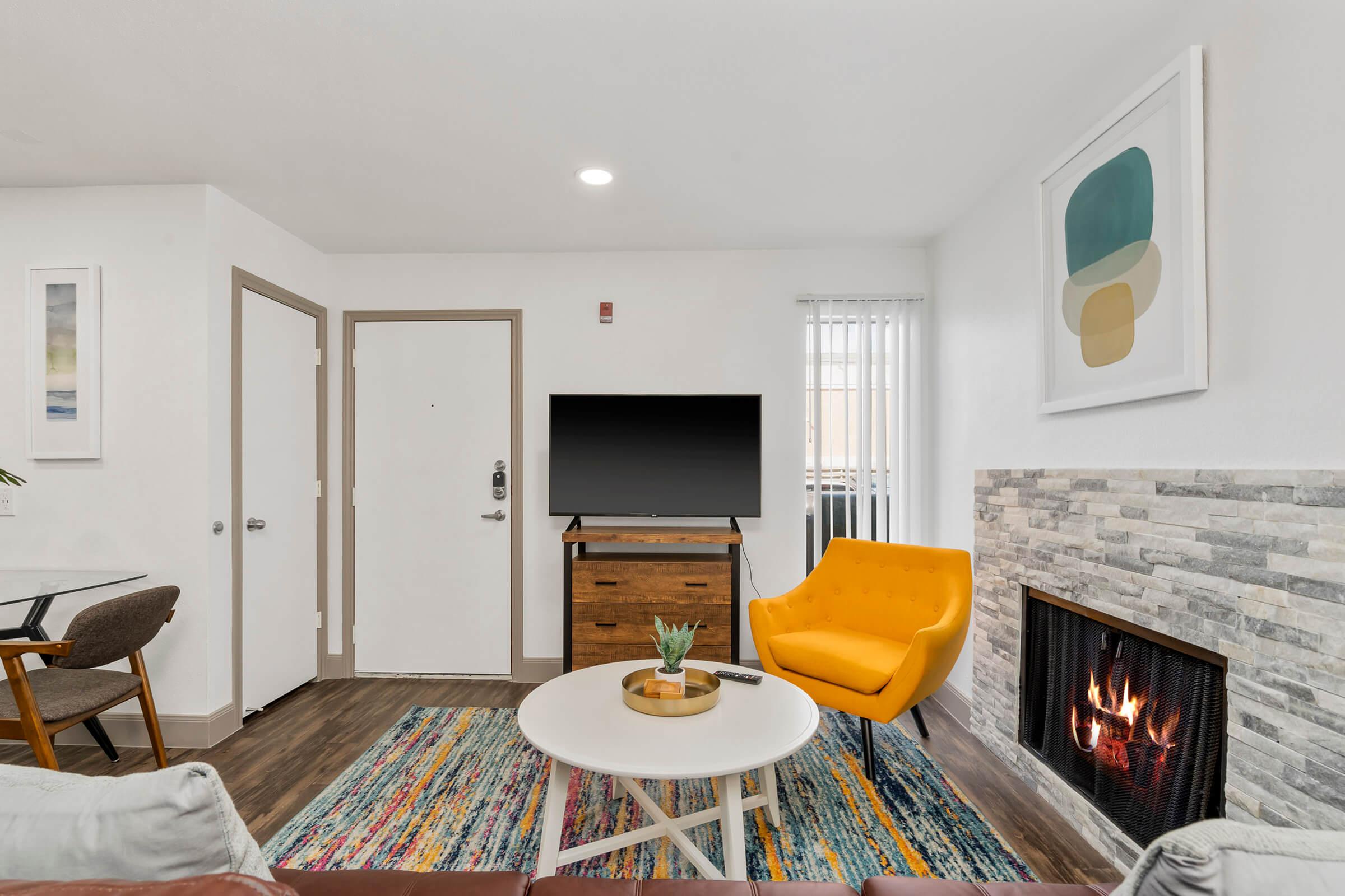 A cozy living room featuring a bright yellow armchair, a round coffee table, and a television on a wooden stand. The space includes a decorative fireplace with flames, a colorful area rug, and modern artwork on the walls. Natural light comes through vertical blinds on a window.