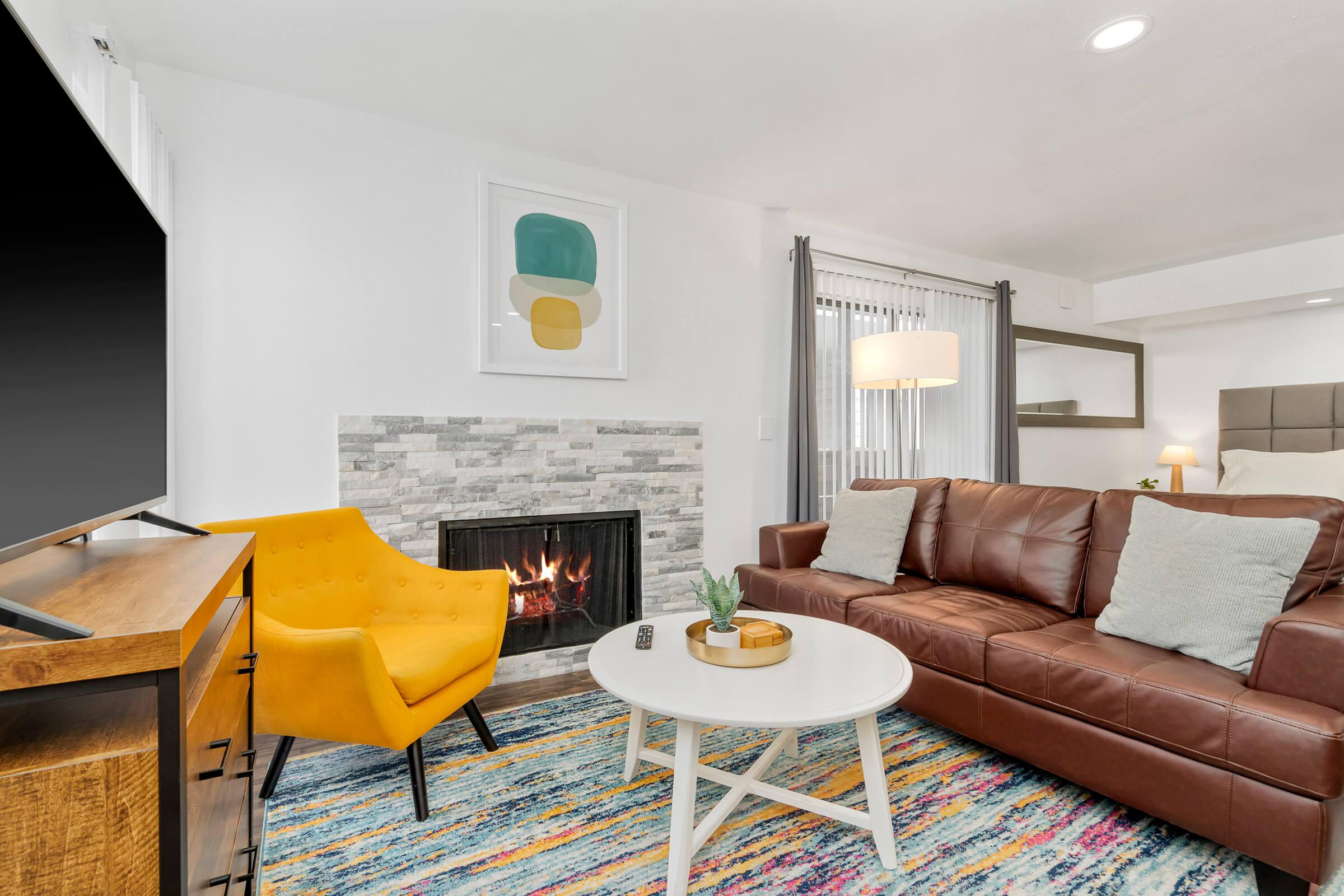 A modern living room featuring a brown leather sofa, a vibrant yellow accent chair, and a round white coffee table. A stone fireplace adds warmth to the space, while colorful area rugs enhance the decor. Large windows with sheer curtains let in natural light, creating a cozy atmosphere.