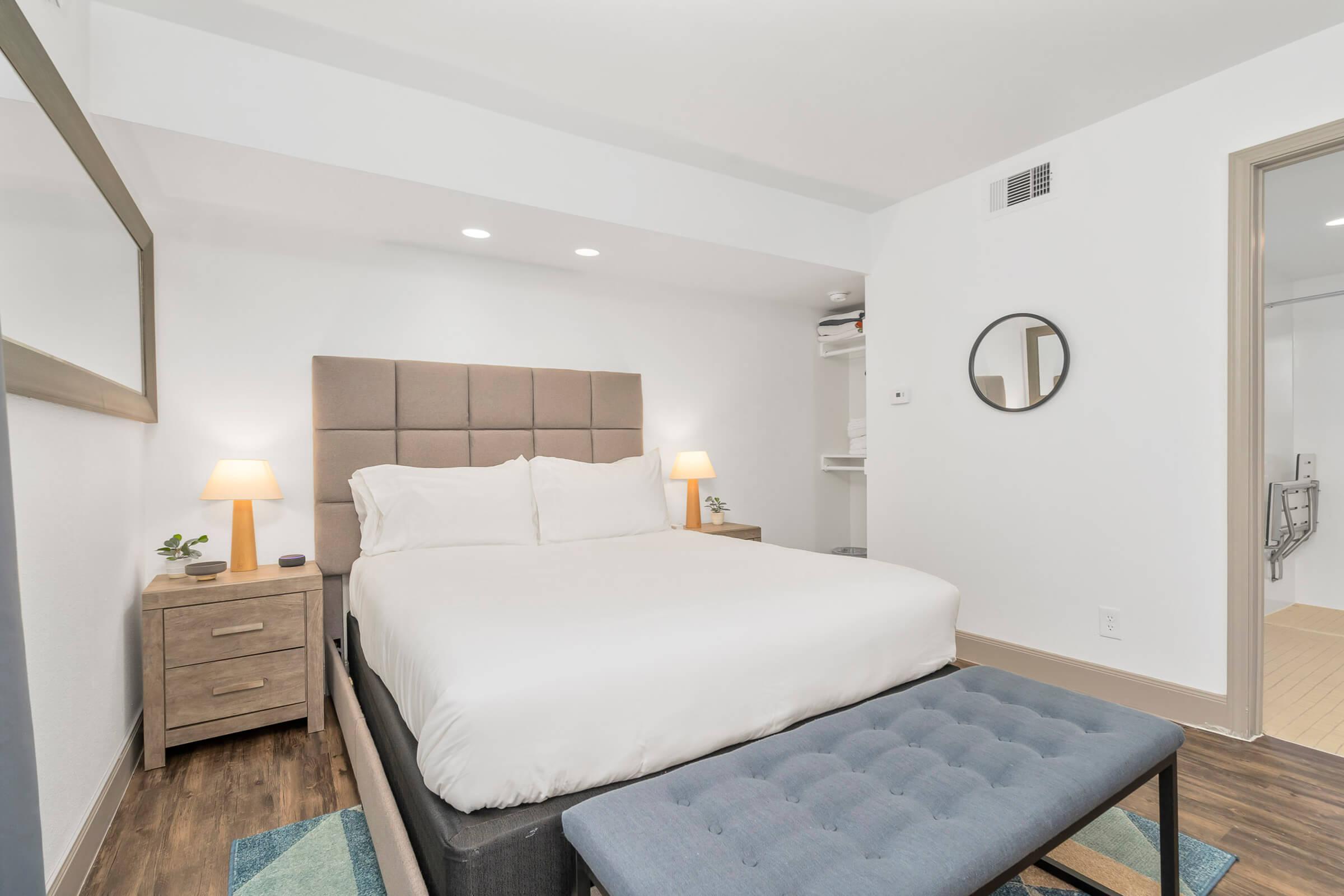 A modern bedroom featuring a queen-sized bed with white bedding, two bedside lamps on wooden nightstands, and a gray upholstered bench at the foot of the bed. The room has light wooden flooring, a large mirror on one wall, and a doorway leading to a bathroom. Soft lighting creates a cozy atmosphere.