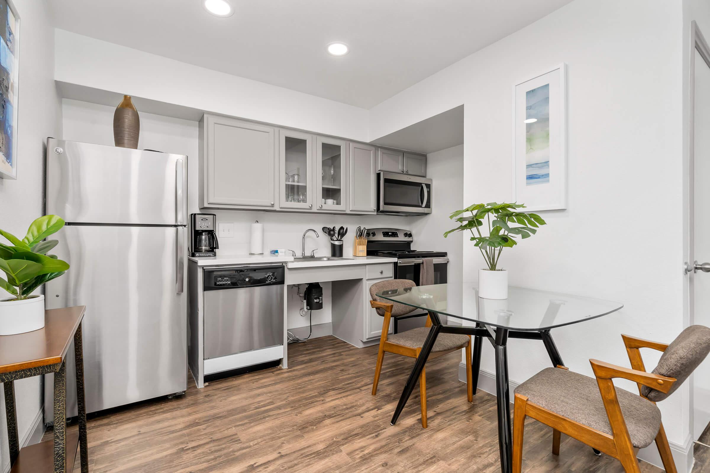 Modern kitchen featuring stainless steel appliances including a fridge, dishwasher, and microwave. There is a glass dining table with wooden chairs, and a potted plant adds a touch of greenery. The walls are painted in a light color, creating a bright and airy atmosphere.