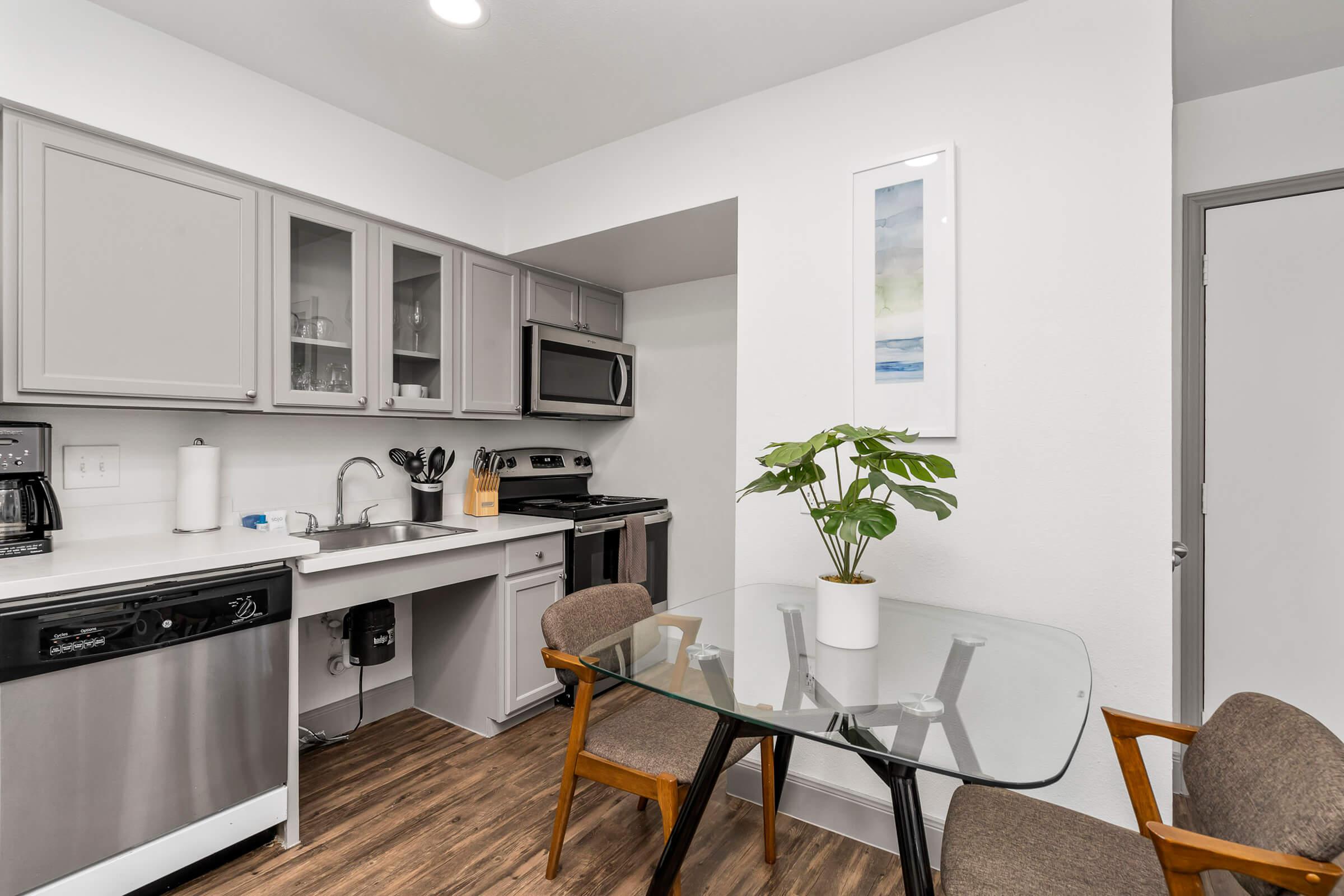 A modern kitchen featuring gray cabinets, stainless steel appliances, and a glass dining table with two wooden chairs. A potted plant sits on the table, and a piece of artwork is displayed on the wall. The space has a clean, minimalist aesthetic with wood-like flooring.