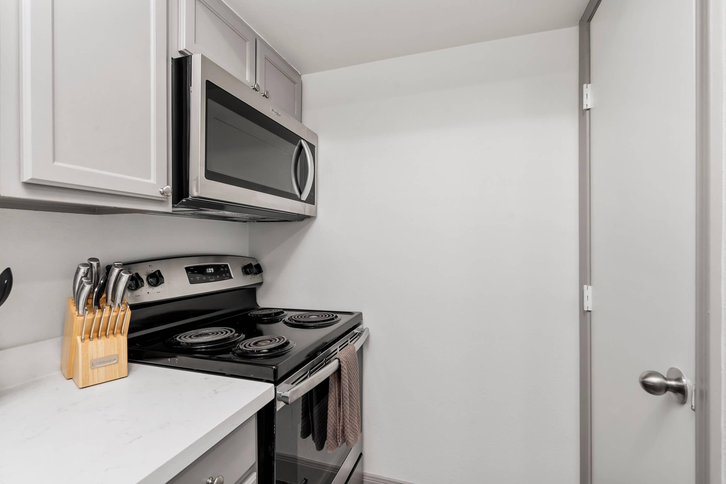 A modern kitchen corner featuring a stainless steel stove and microwave above it. There is a white countertop with a wooden knife block. The wall is painted white, and a door is partially visible on the right. The overall decor is sleek and minimalist.