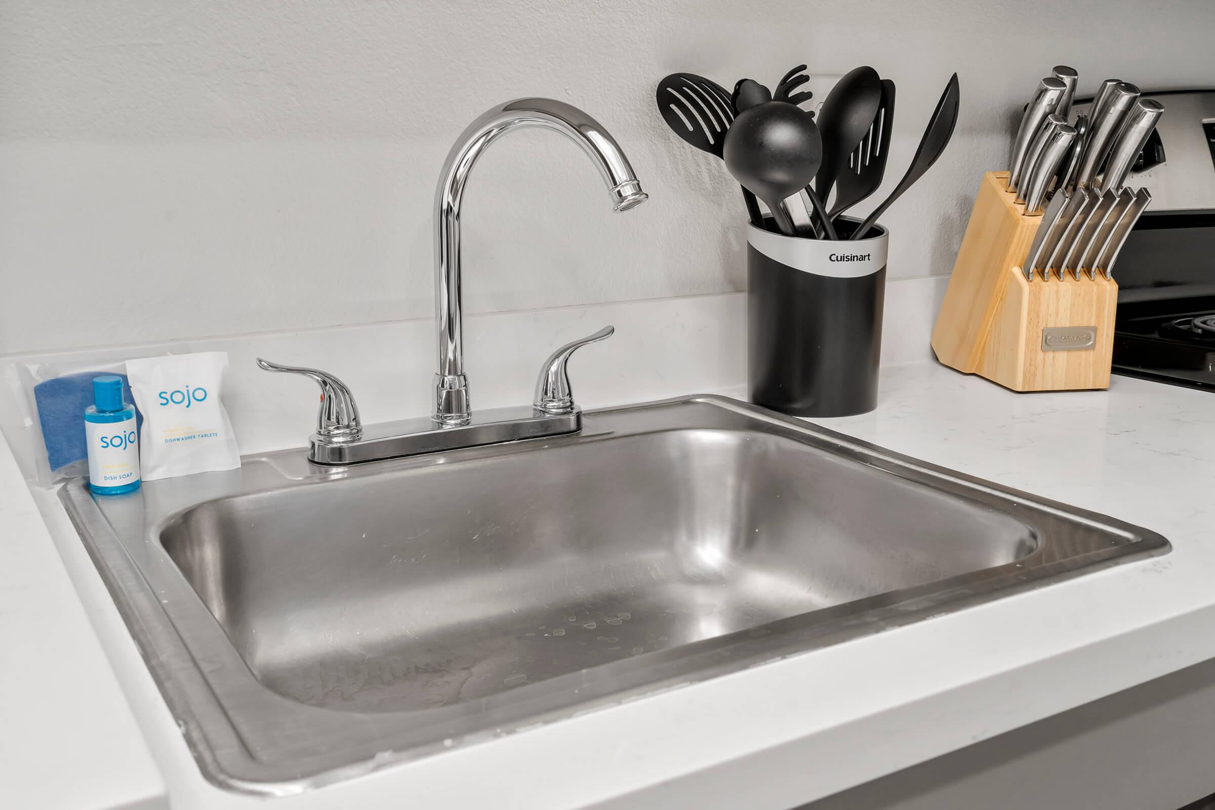 A stainless steel sink with a polished chrome faucet sits on a white countertop. Next to the sink, there is a small blue bottle labeled "sojo" and a folded napkin. A black utensil holder with kitchen tools and a wooden knife block with various knives are also visible in the background.