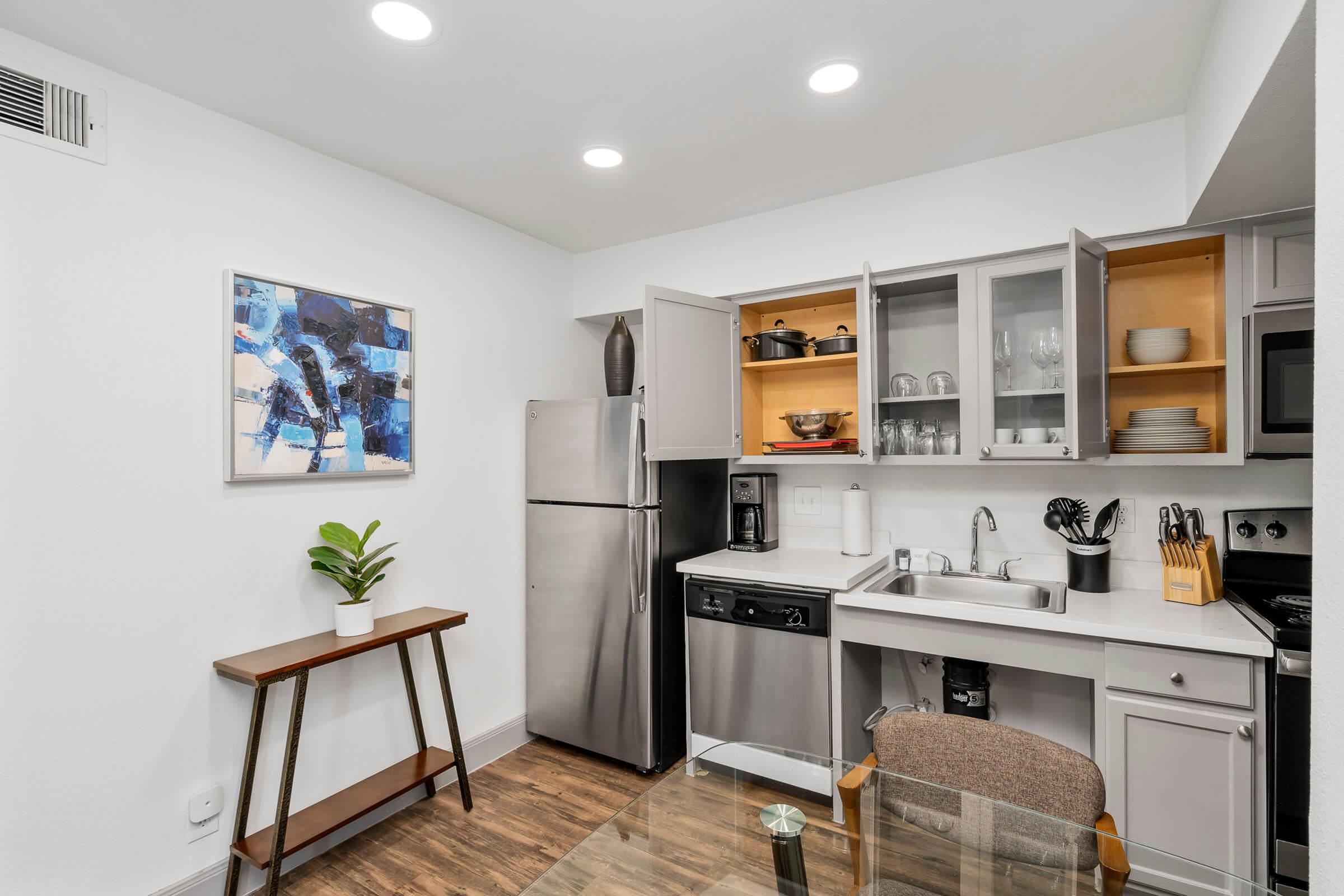 A modern kitchen featuring a stainless steel refrigerator, microwave, and dishwasher. Open shelves display dinnerware, and a small table with a plant adds a touch of greenery. The space has neutral-colored walls and wooden flooring, creating a bright and inviting atmosphere.