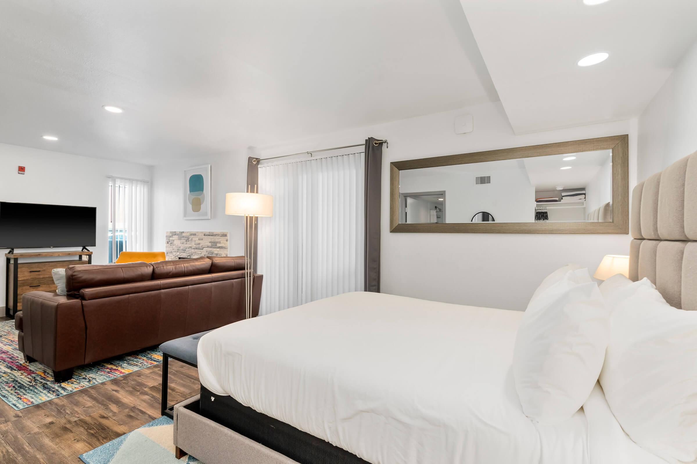 A spacious hotel room featuring a king-sized bed with white linens, a brown leather sofa, a flat-screen TV, and a stylish lamp. Natural light fills the space through large windows with sheer curtains, highlighting the modern decor and colorful area rug. A mirror adds depth to the room's design.