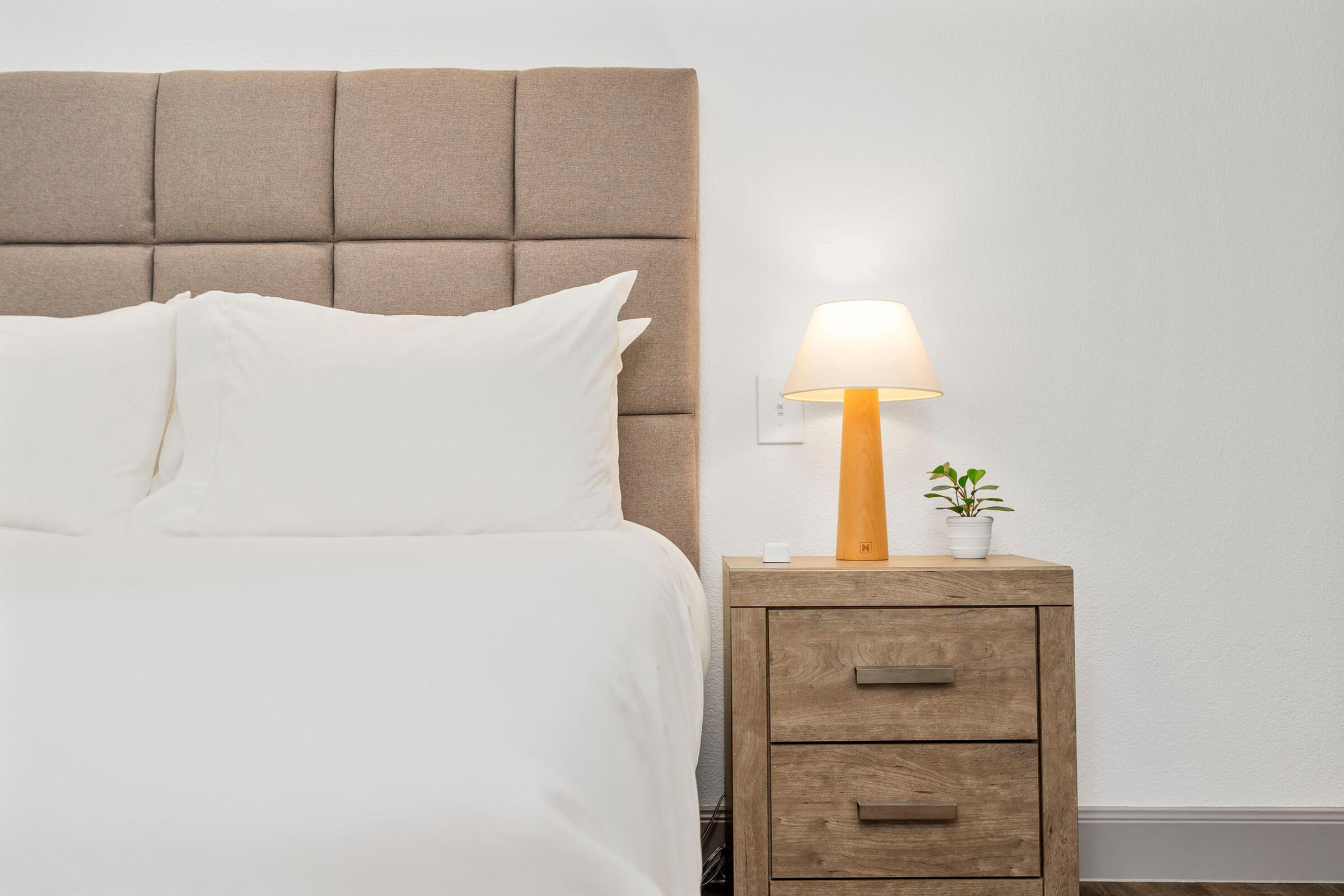 A neatly made bed with white pillows and a beige upholstered headboard. Next to the bed is a wooden nightstand with two drawers, featuring a small potted plant and a warm-lit lamp on top. The background wall is a clean, light color, creating a calm and modern bedroom atmosphere.