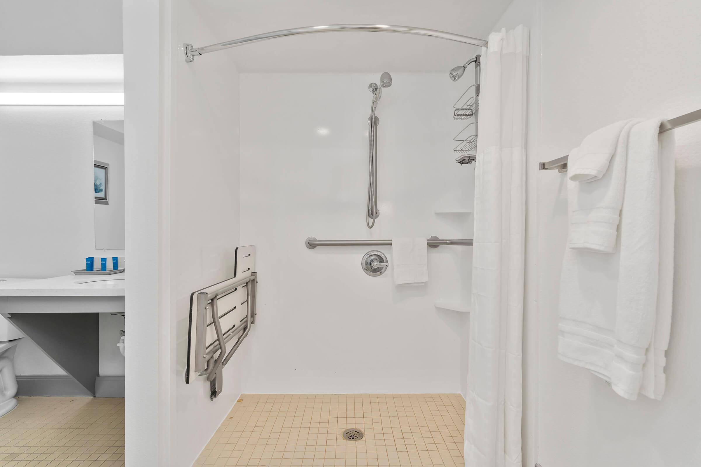 A clean, accessible bathroom with a walk-in shower featuring grab bars, a handheld showerhead, and shower shelves. Two white towels hang on a towel bar, and a small corner shelf is visible. In the background, a sink area with toiletries can be seen.