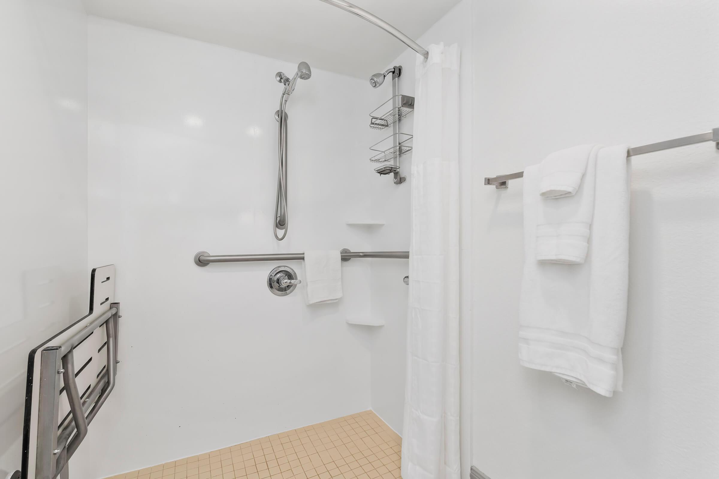 A clean, accessible bathroom shower stall featuring a curved shower rod, a white shower curtain, and grab bars. Two neatly folded white towels hang on a towel rack, and there are shelves for toiletries. The floor is tiled in a light color, creating a bright and inviting atmosphere.