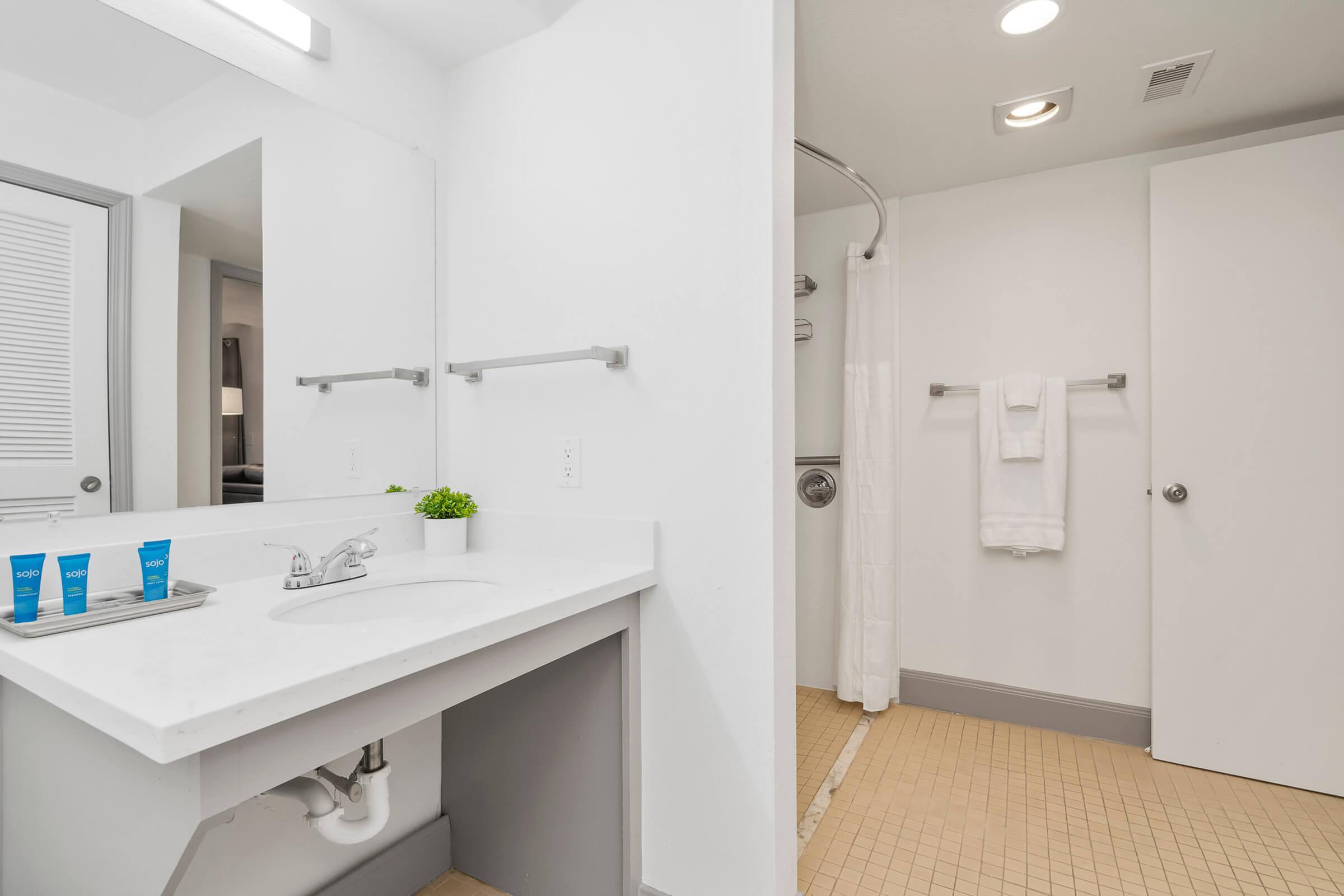 A modern bathroom featuring a white sink with a countertop, two blue toiletries placed on a tray, a mirror, and a shower area with a glass door. Soft lighting enhances the clean design, while beige tiled flooring contrasts with the white walls. A towel rack with neatly hung white towels is visible.