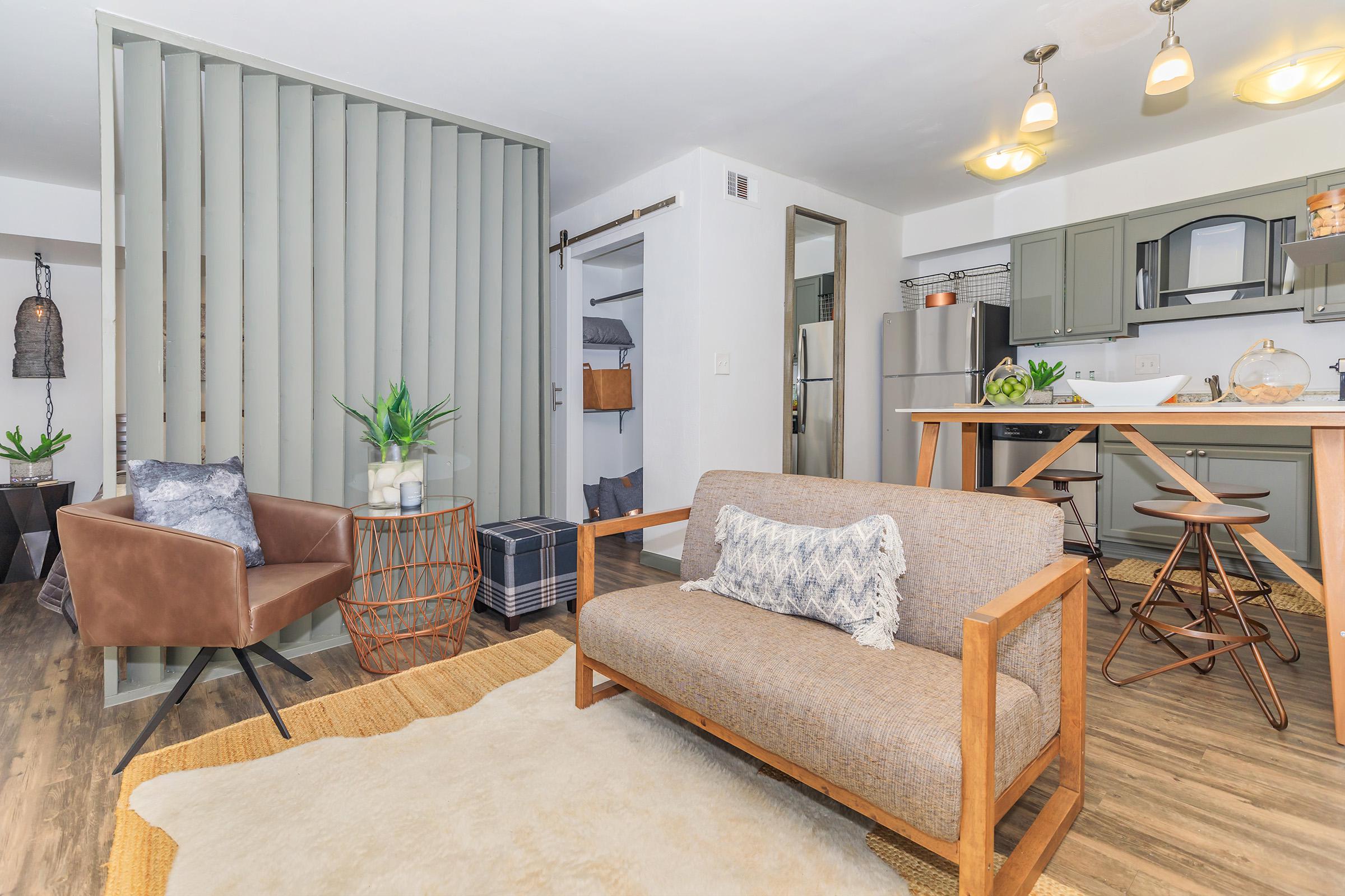 A cozy living area featuring a modern sofa, a brown accent chair, and a round table with plants. The space has an open layout with a kitchen in the background, showcasing gray cabinets and stylish light fixtures. A decorative rug covers the floor, adding warmth to the contemporary decor.