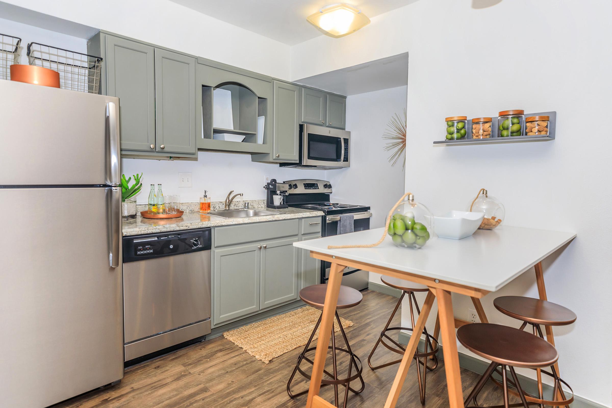 A modern kitchen featuring gray cabinets, stainless steel appliances, and a countertop with bar stools. A small dining table sits in the foreground. The kitchen has a decorative touch with plants and a bowl of fruit, complemented by natural light and a warm, welcoming atmosphere.