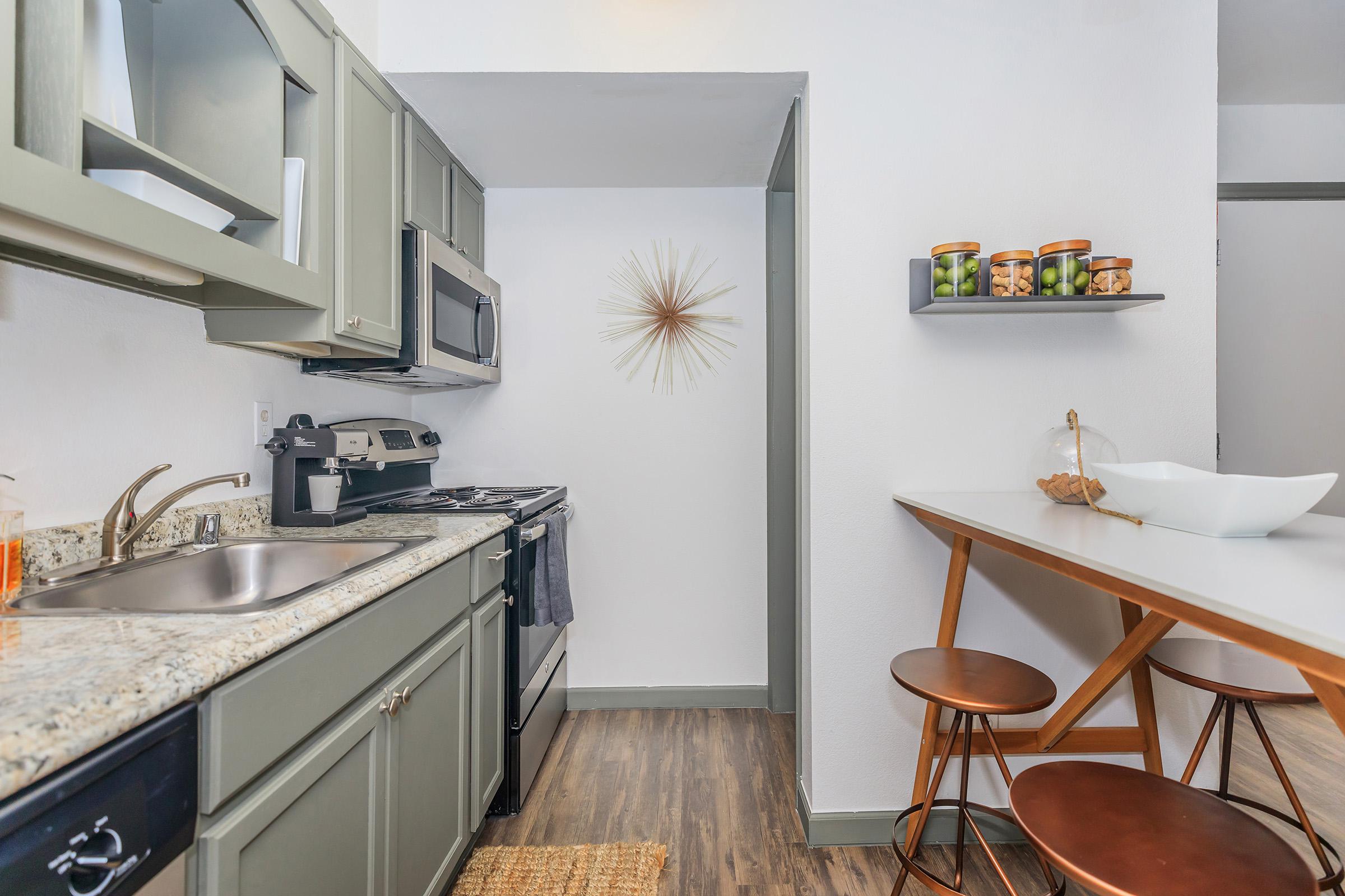 A modern kitchen featuring olive green cabinets, stainless steel appliances, and a granite countertop. The space includes a small dining area with a minimalist table and copper stools. Decorative items, including a starburst wall clock and jars of pickles, add a stylish touch to the bright and inviting atmosphere.