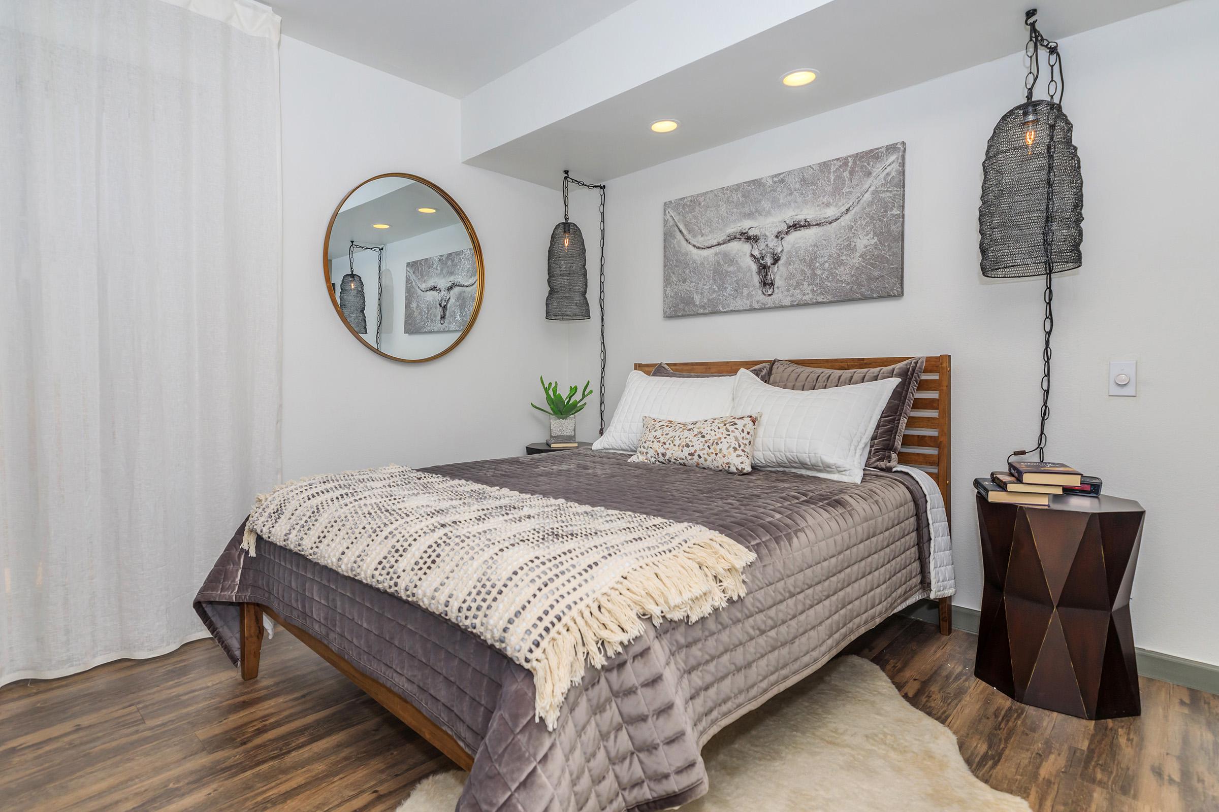 A cozy bedroom featuring a wooden bed with a gray blanket and decorative pillows. The walls are adorned with a circular mirror and a rustic artwork of a bull's head. Soft lighting is provided by pendant lamps, and a small plant is on the bedside table, with books and a decorative accent beside it. A light rug completes the space.