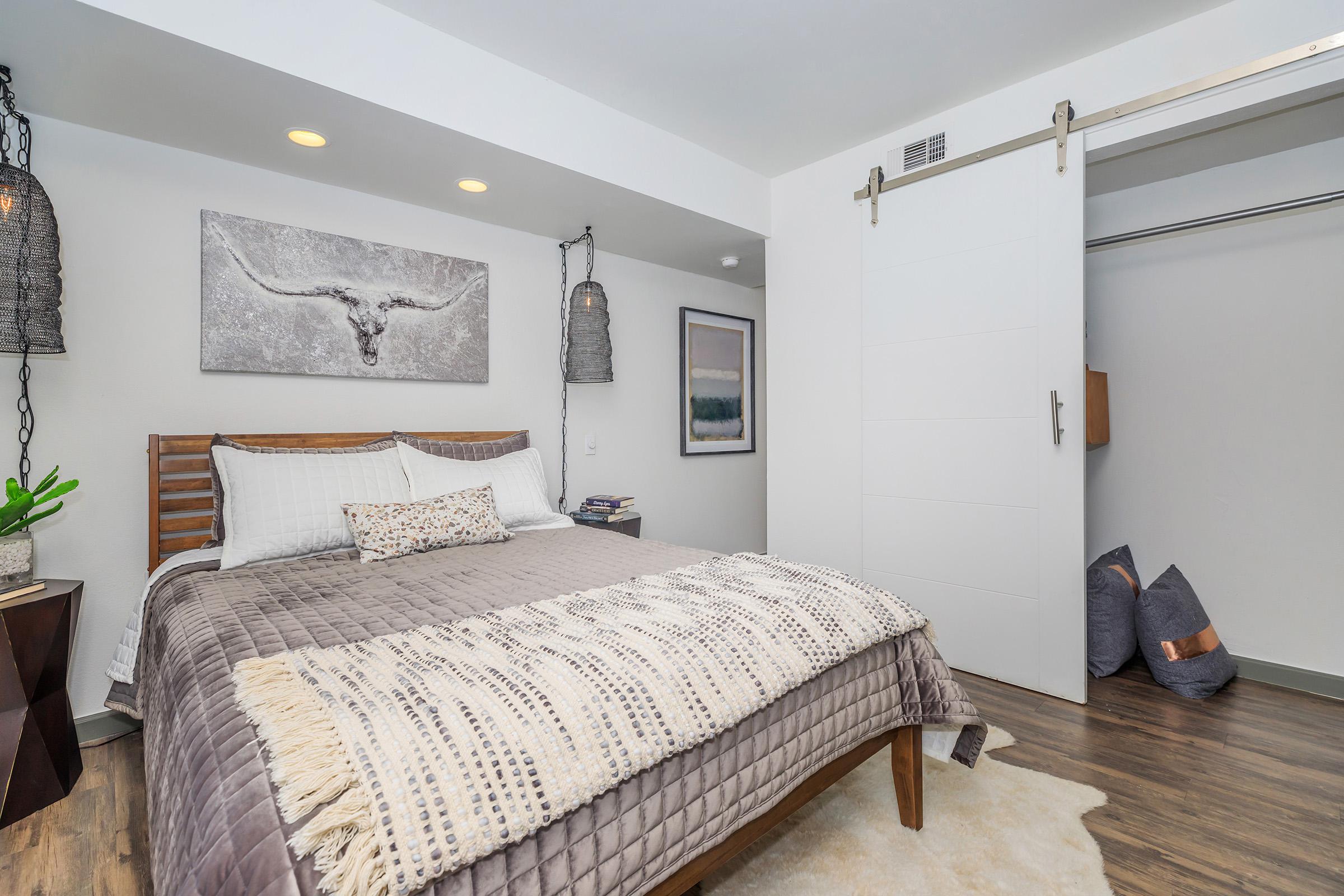 A modern bedroom featuring a wooden bed frame with a gray blanket and decorative pillows, a wall art piece depicting a bull skull, two stylish pendant lights, and a sliding white closet door. The floor is wood, and there are accent pillows in the corner. A potted plant adds a touch of greenery.