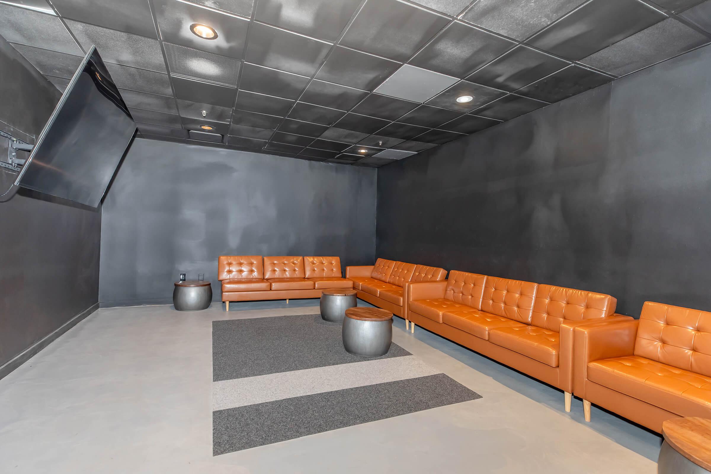 Modern lounge area featuring sleek orange leather couches arranged around a circular metal coffee table. A large flat-screen TV is mounted on the black wall, and a gray striped rug lies on the floor, contributing to a minimalist and stylish atmosphere.