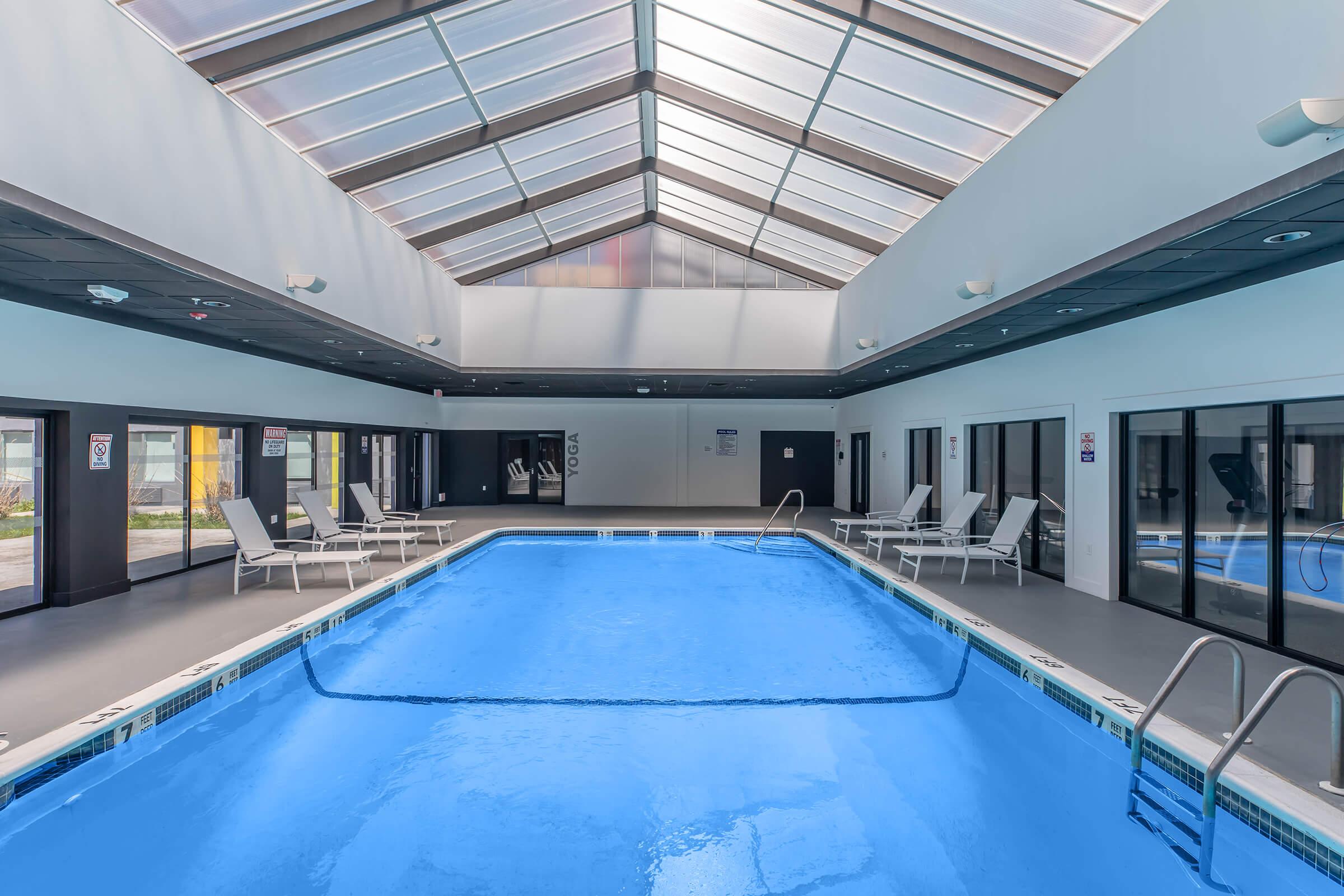 Indoor swimming pool with a clear, blue water surface. Surrounded by lounge chairs, the pool area features large windows and a skylight, providing natural light. The walls are painted in neutral tones, and there are doors leading to the outside. The atmosphere is bright and inviting.