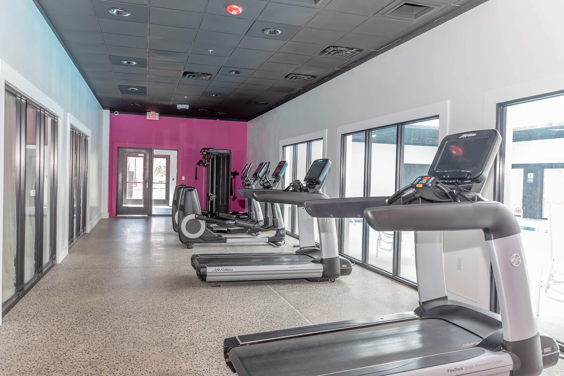 A modern gym interior featuring several treadmills and exercise equipment lined up against large windows. The walls are painted in a combination of light and vibrant pink, creating a bright and inviting atmosphere. The flooring is a smooth, polished surface, adding to the contemporary design.