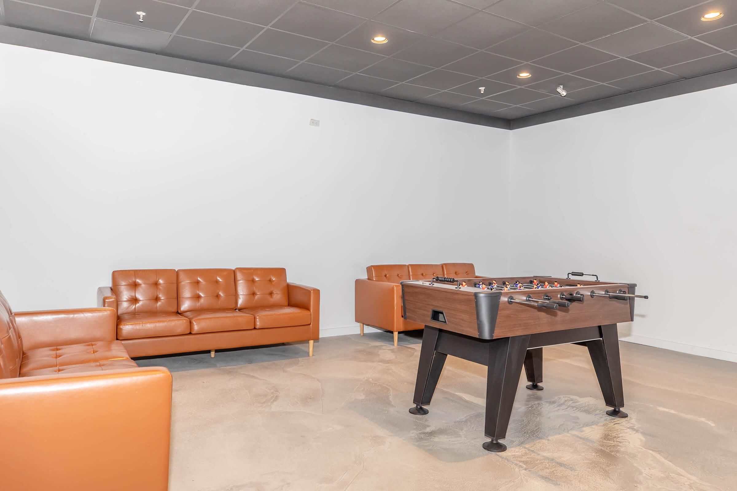 A cozy lounge area featuring two brown leather sofas and a foosball table on a polished concrete floor. The walls are white, and the ceiling has recessed lighting, creating a minimalist and inviting space for relaxation or casual games.