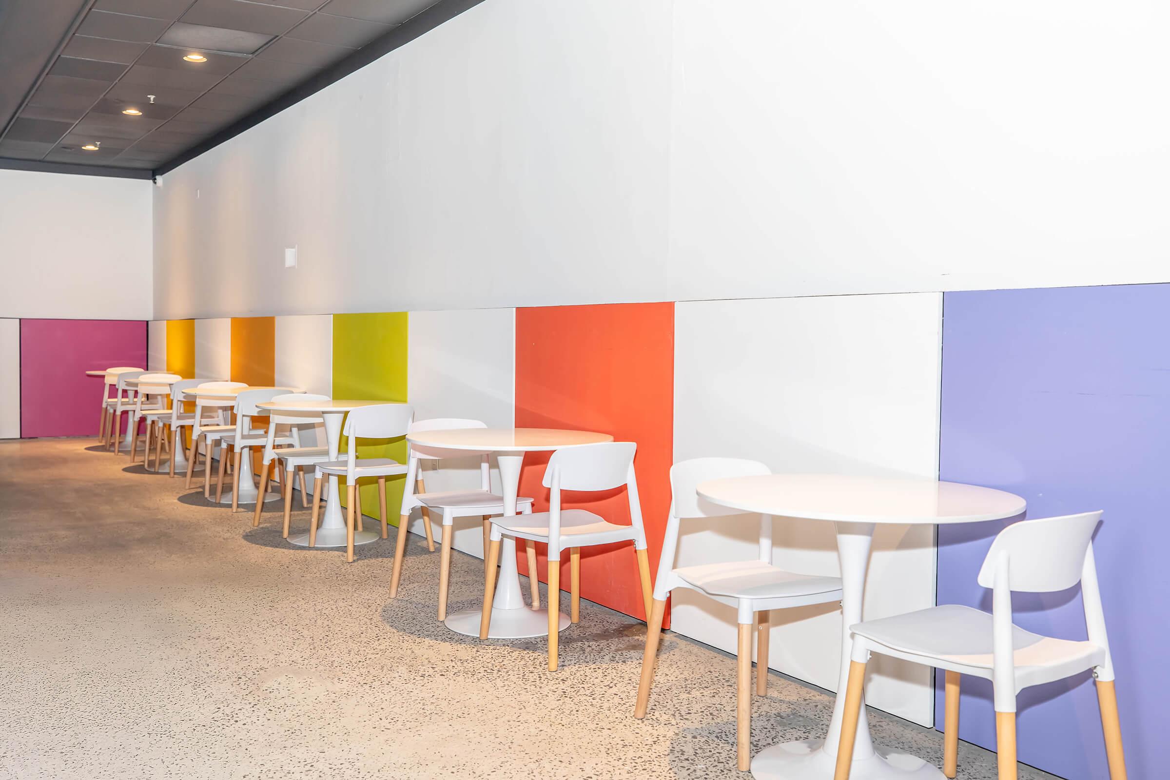 A modern café interior featuring white tables and chairs arranged along a wall. The wall is painted in vibrant colors, including orange, purple, and yellow, creating a lively atmosphere. The flooring is textured concrete, adding to the contemporary design of the space.