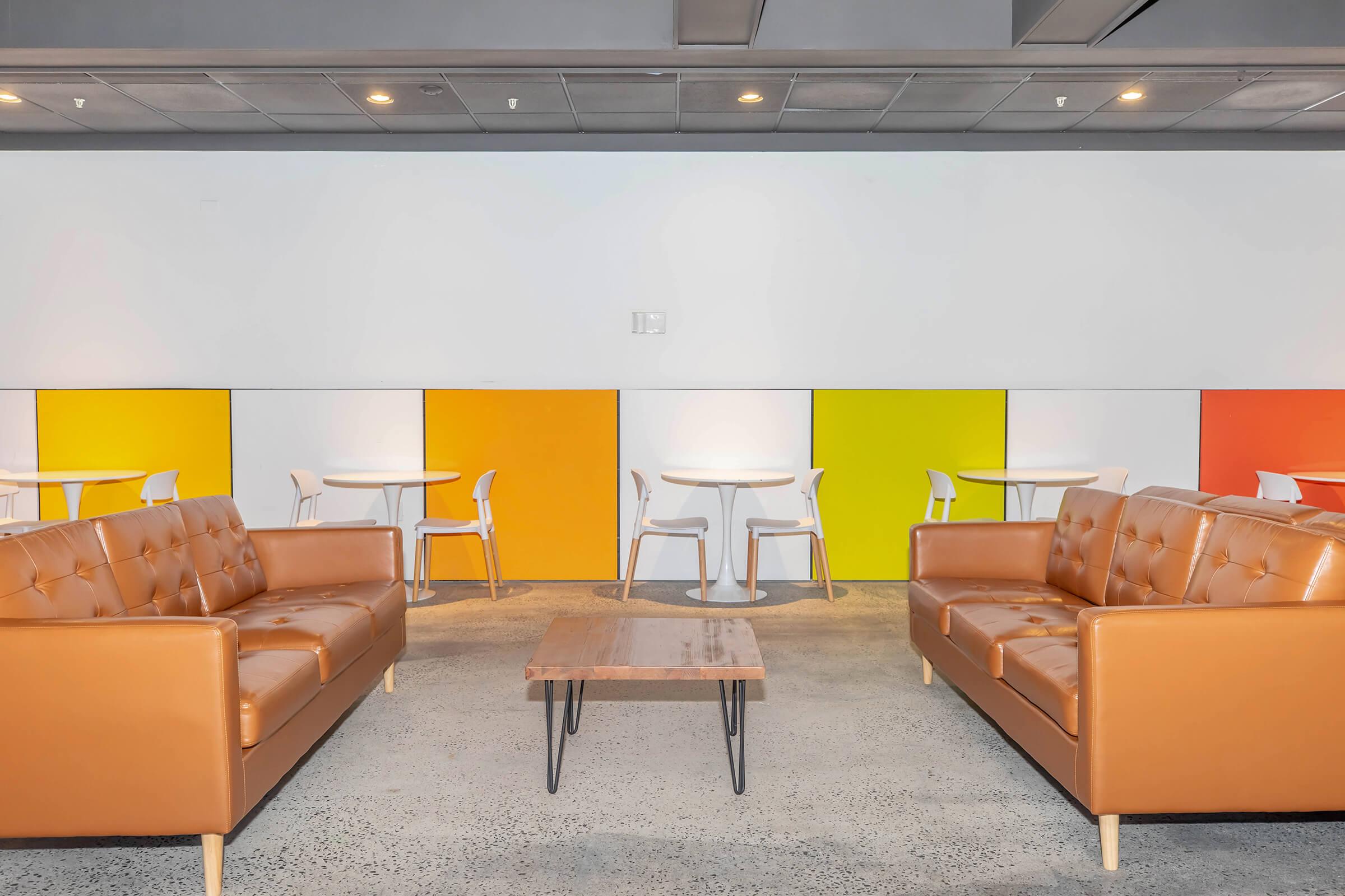 A modern lounge area featuring two brown leather sofas facing a wooden coffee table, and colorful wall panels in orange, yellow, and white. In the background, there are several round tables with white chairs, creating an inviting and trendy atmosphere.