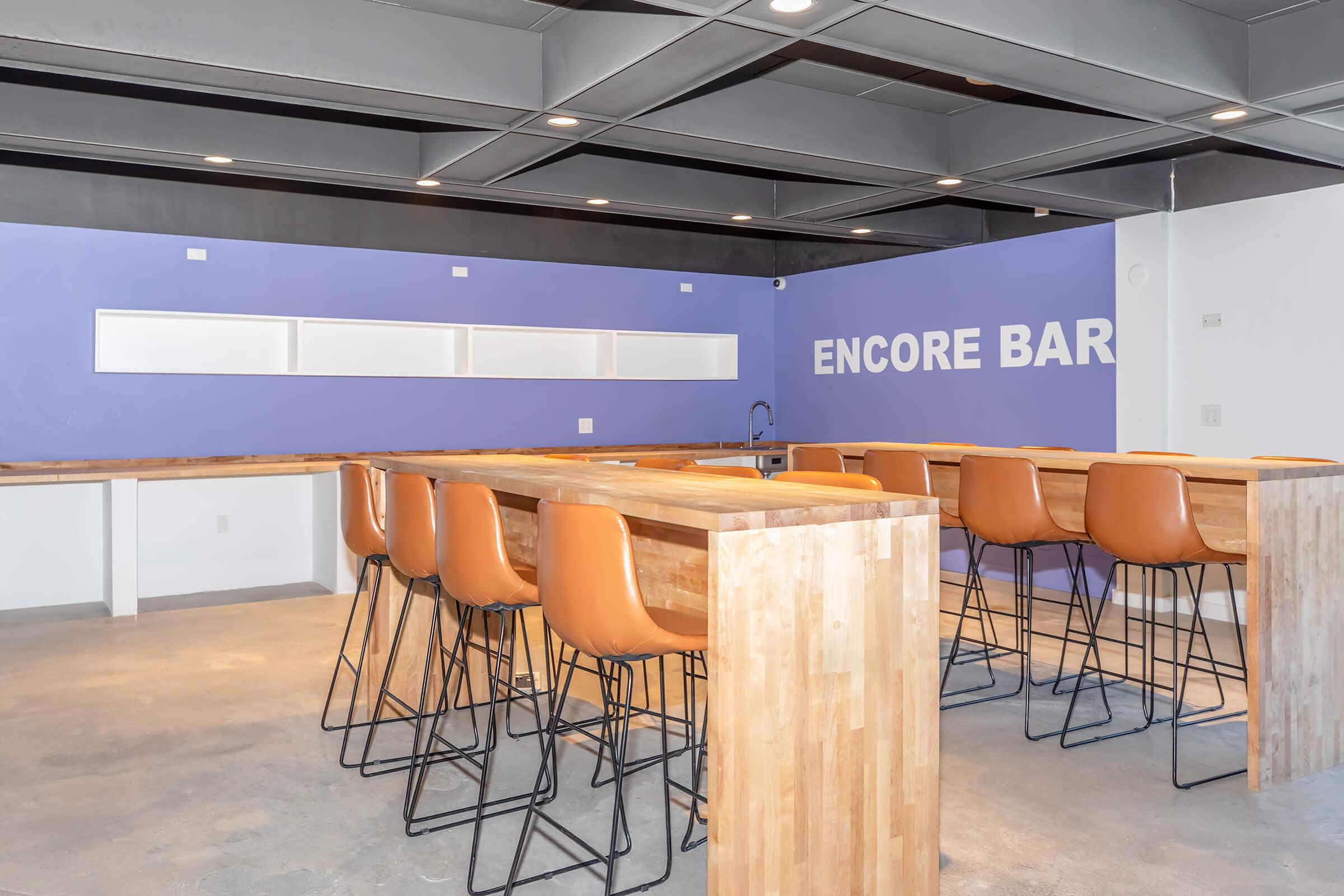 A modern interior of a space featuring a bar with wooden countertops and high stools with orange upholstery. The wall behind is painted purple with the text "ENCORE BAR" displayed prominently. The atmosphere is contemporary and inviting, suitable for social gatherings or events.