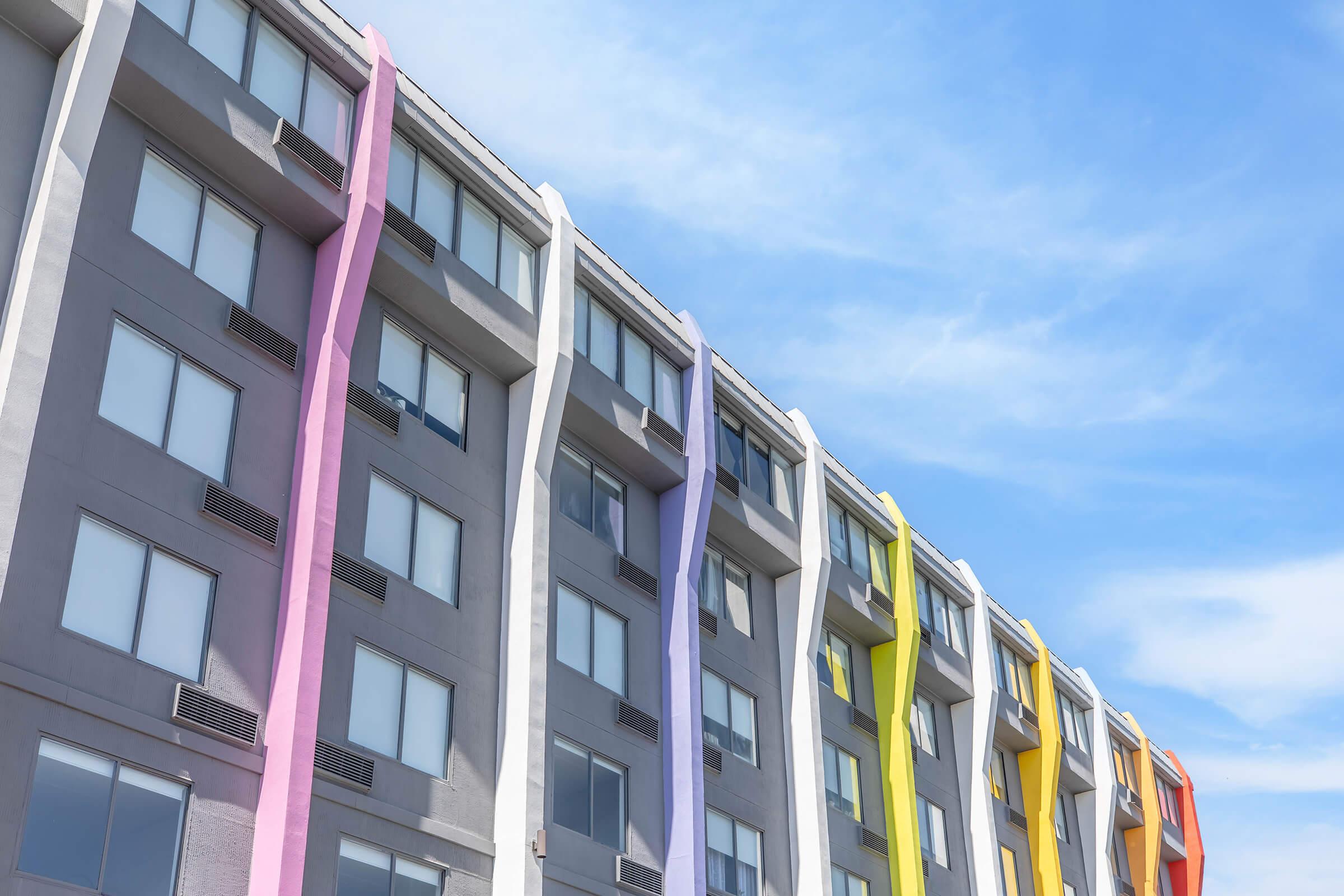 A modern building featuring a gray facade with colorful vertical accents in pink, lavender, and yellow. The structure has multiple windows, and the vibrant colors contrast against the blue sky, creating a lively and inviting atmosphere.