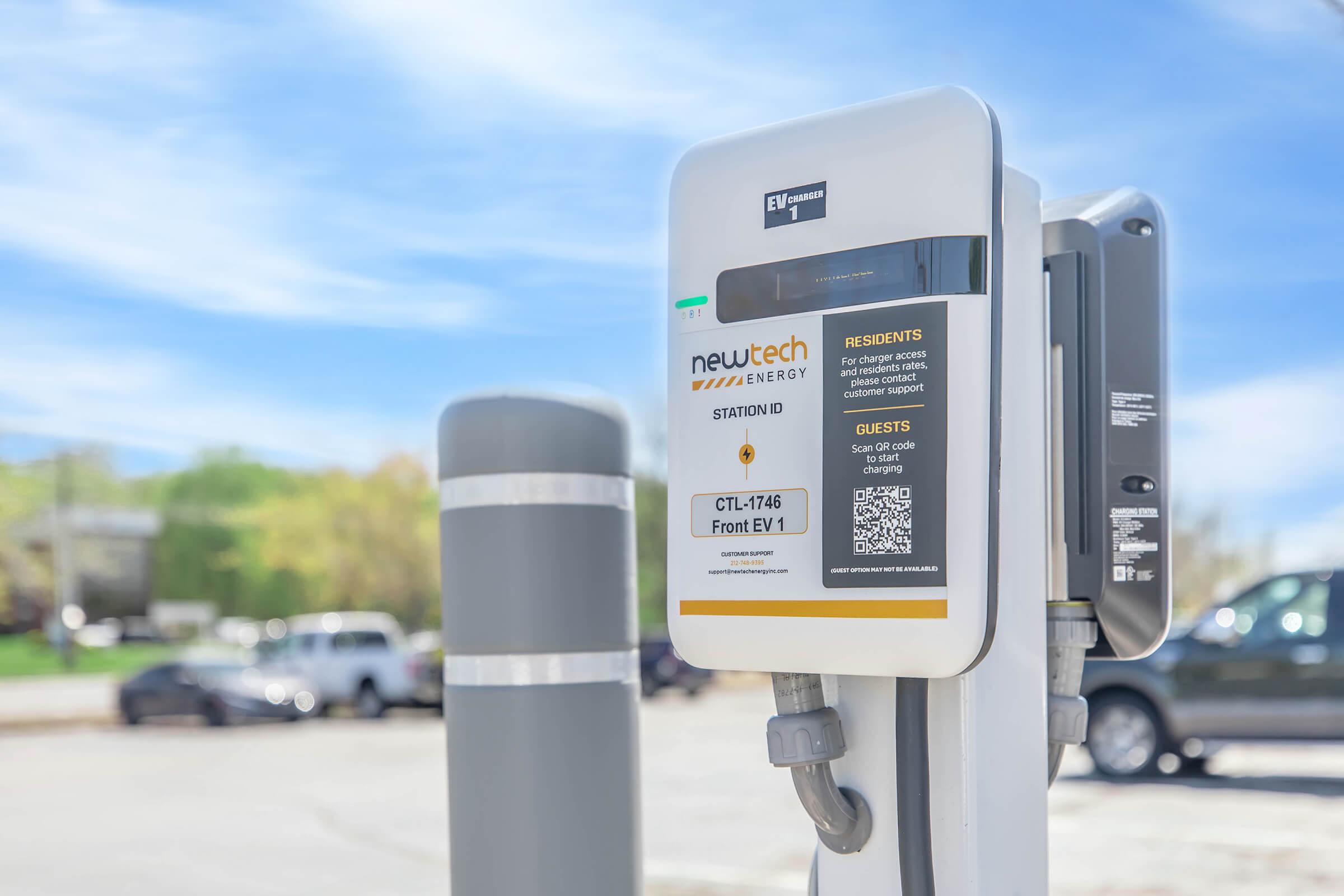 Electric vehicle charging station labeled "newtech ENERGY," featuring a QR code for access. The station is designed for residents and guests, with instructions visible on a screen. The background includes a clear blue sky and vehicles parked nearby.