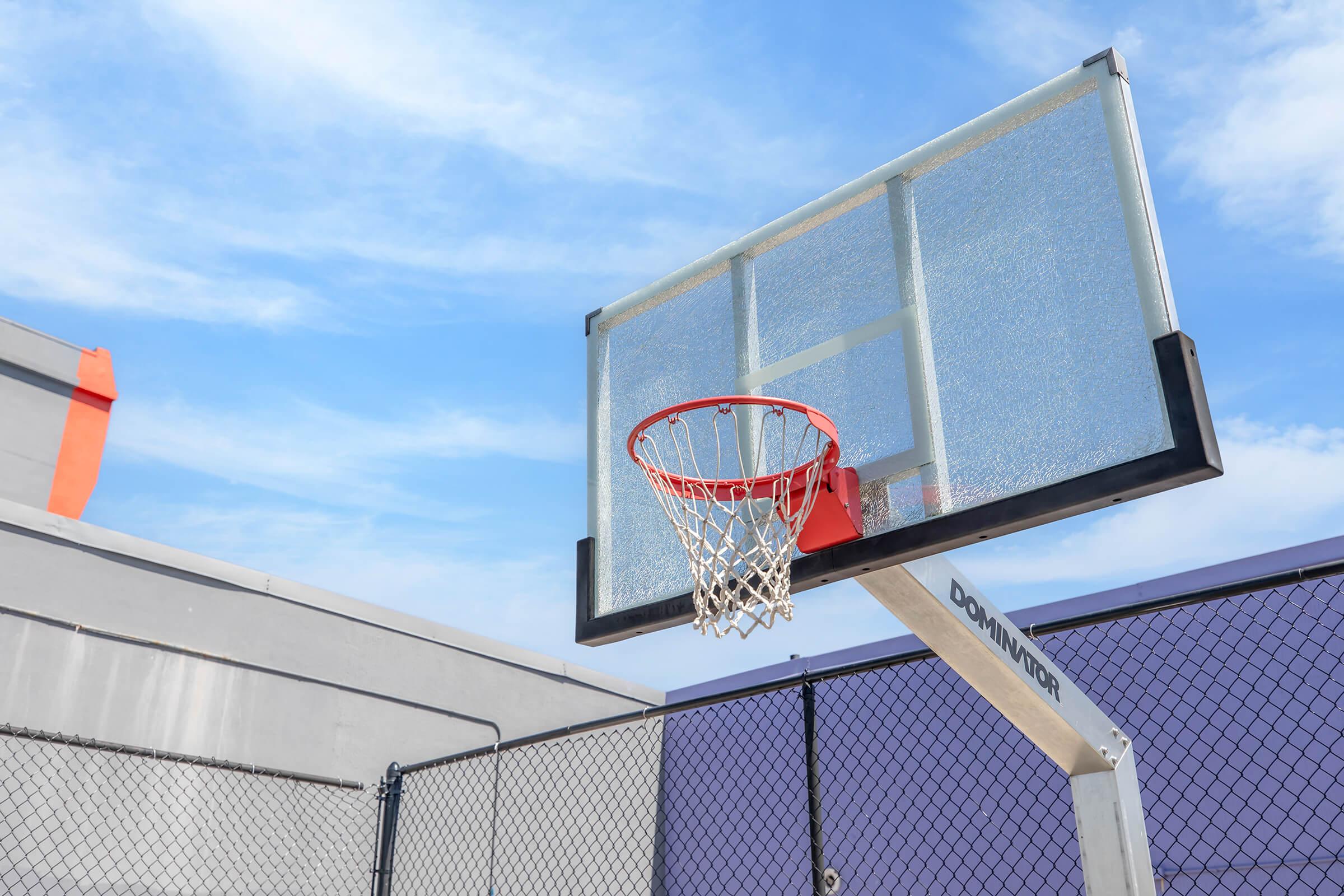 A basketball hoop mounted on a pole, with a clear backboard and a red rim. The hoop is situated outdoors with a blue sky and some clouds in the background. A chain-link fence surrounds the area, and a gray building is visible in the distance.