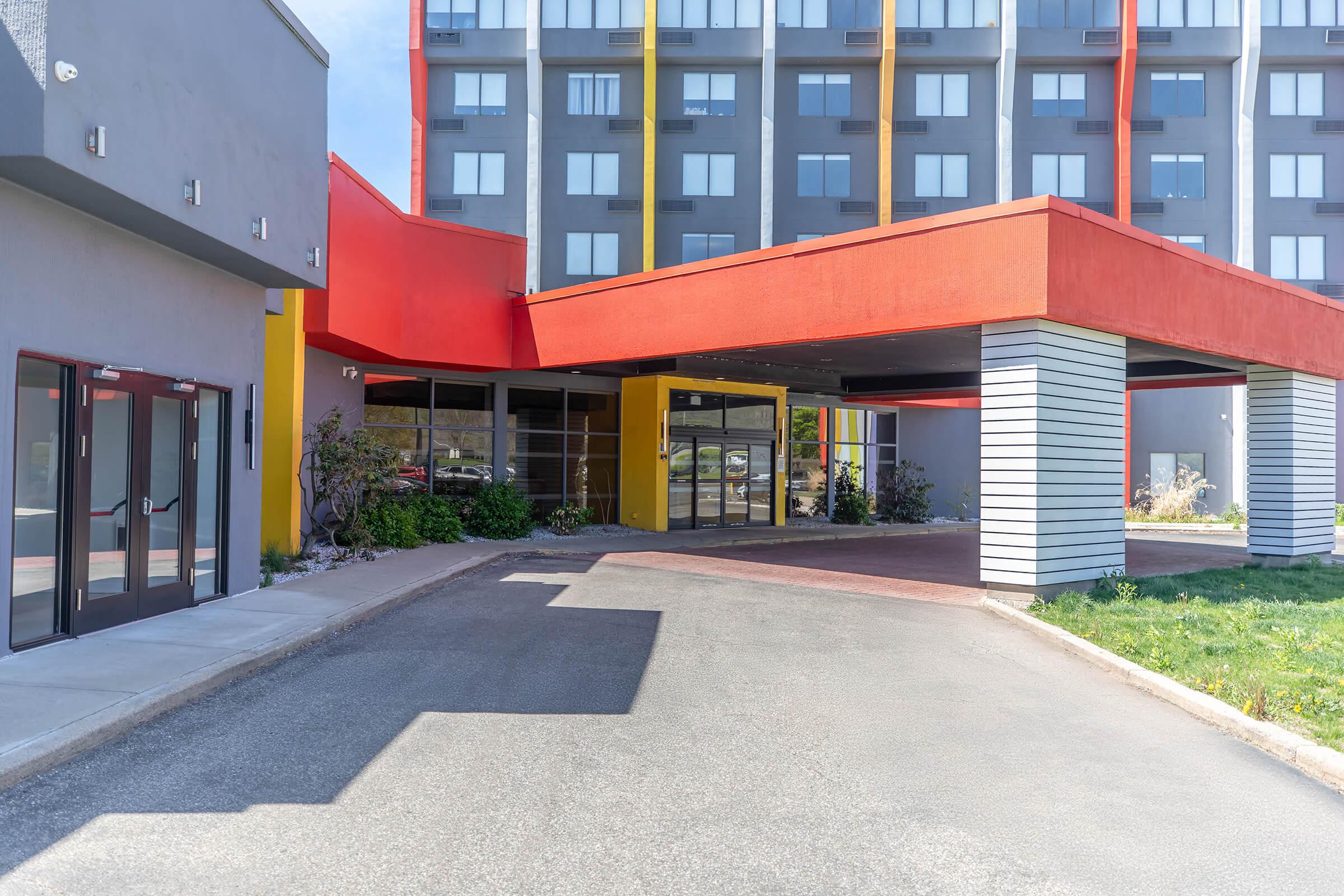 Modern hotel entrance featuring a vibrant red and yellow awning, surrounded by greenery. The building has a contemporary design with large windows and a gray exterior. The pathway leading to the entrance is paved, providing access for guests.