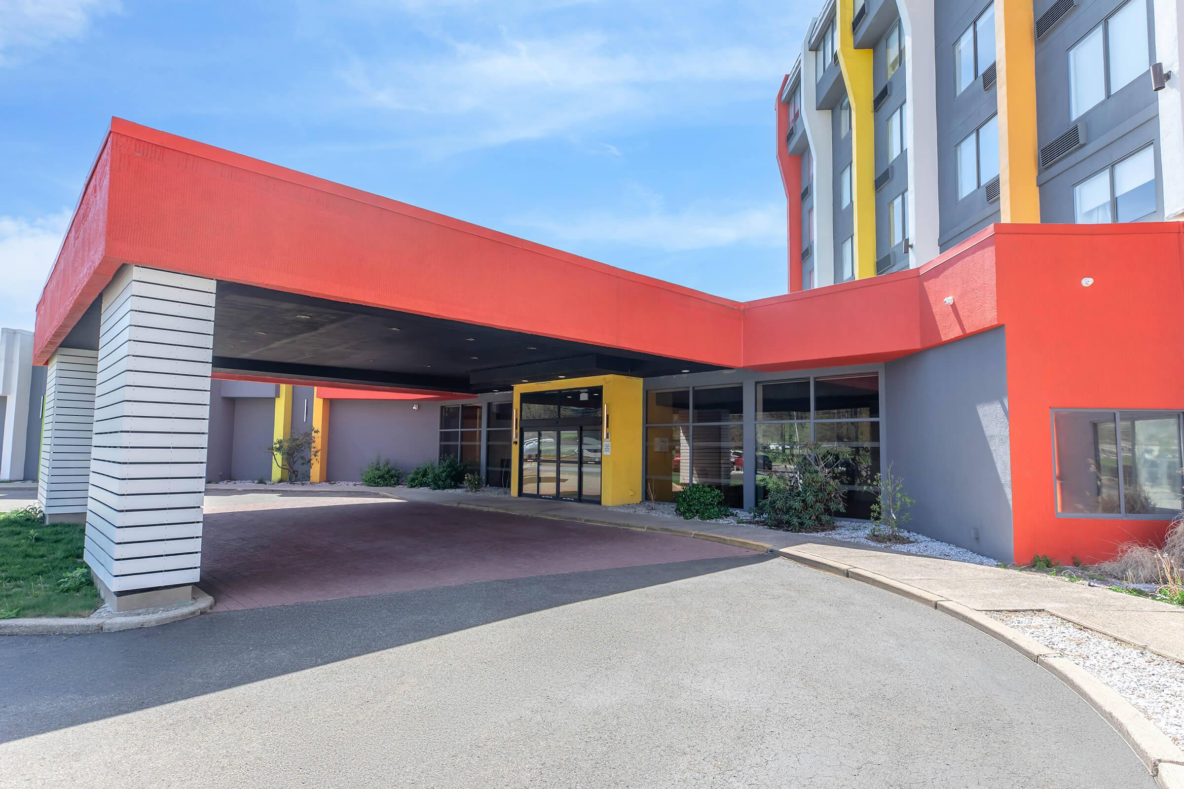 Modern hotel exterior featuring a vibrant red and yellow entrance canopy. The building has a sleek design with large windows and a mix of gray and colorful walls. A paved driveway leads up to the entrance, with landscaped greenery on either side. Clear blue sky above adds to the inviting ambiance.