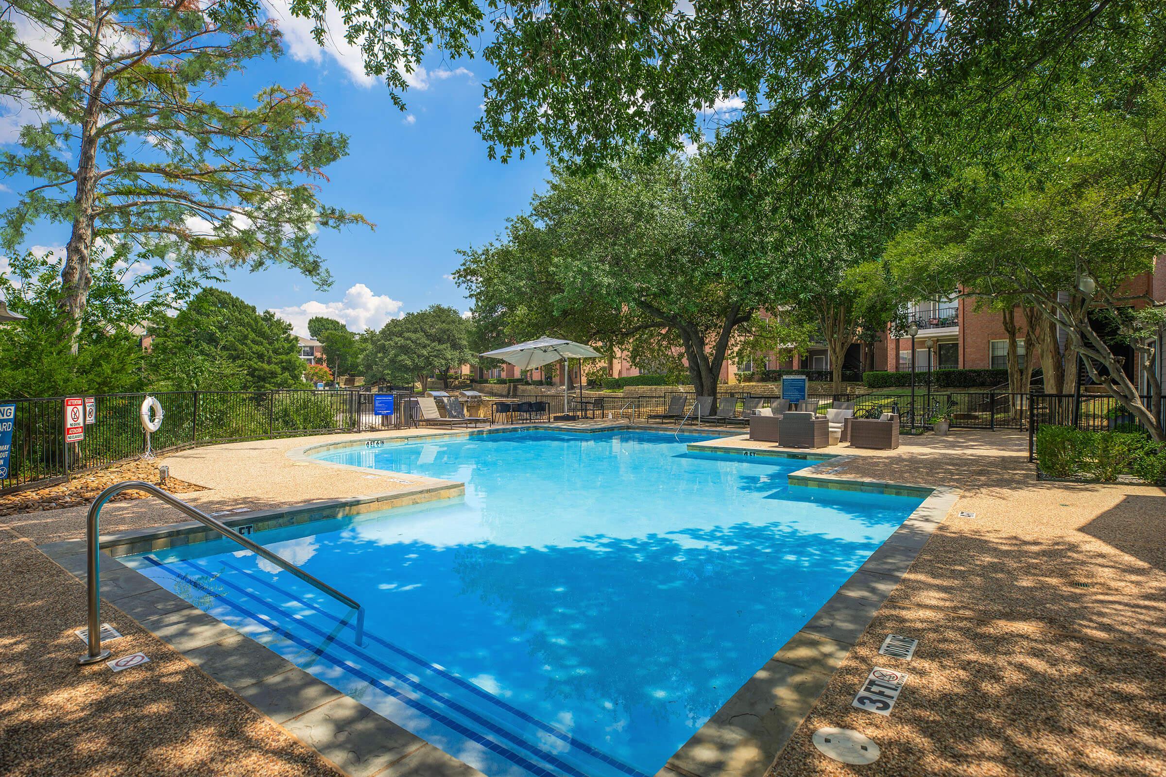 A sunny outdoor swimming pool surrounded by trees and lounge chairs. The pool has a shallow entry with a handrail and is set in a landscaped area with apartments in the background. A shaded seating area is visible, creating a relaxing atmosphere.