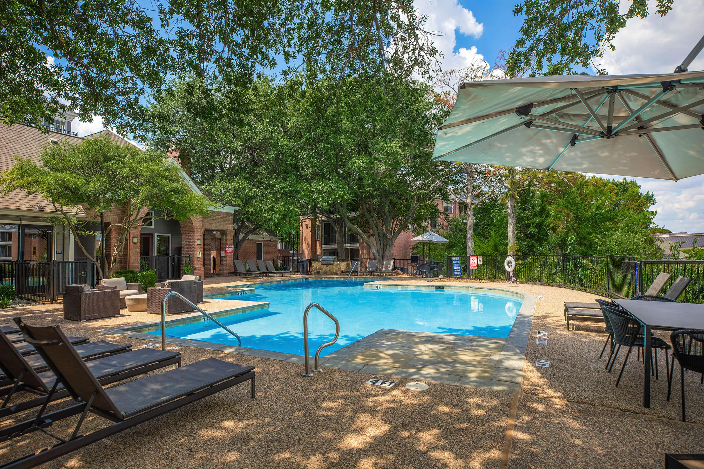 A serene pool area surrounded by lush trees, featuring a clear blue water pool, lounge chairs, and shaded seating. The space is well-lit with a sunny sky above, creating a relaxing atmosphere for outdoor enjoyment.