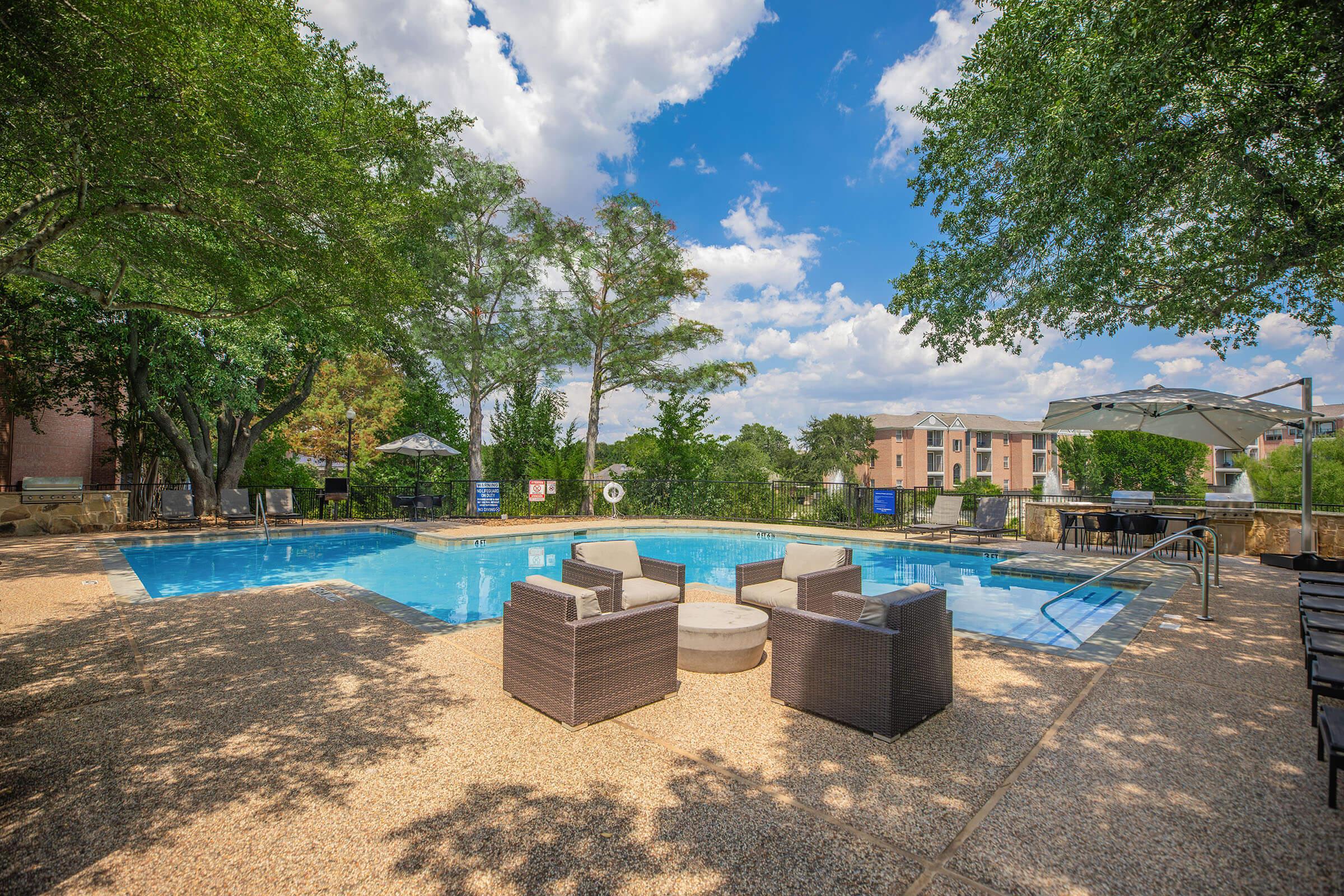 A serene outdoor swimming pool surrounded by lush trees and chairs. The pool area features comfortable seating arrangements, sun loungers, and umbrellas, set against a bright blue sky with fluffy clouds. Nearby buildings are visible in the background, creating a relaxing atmosphere.