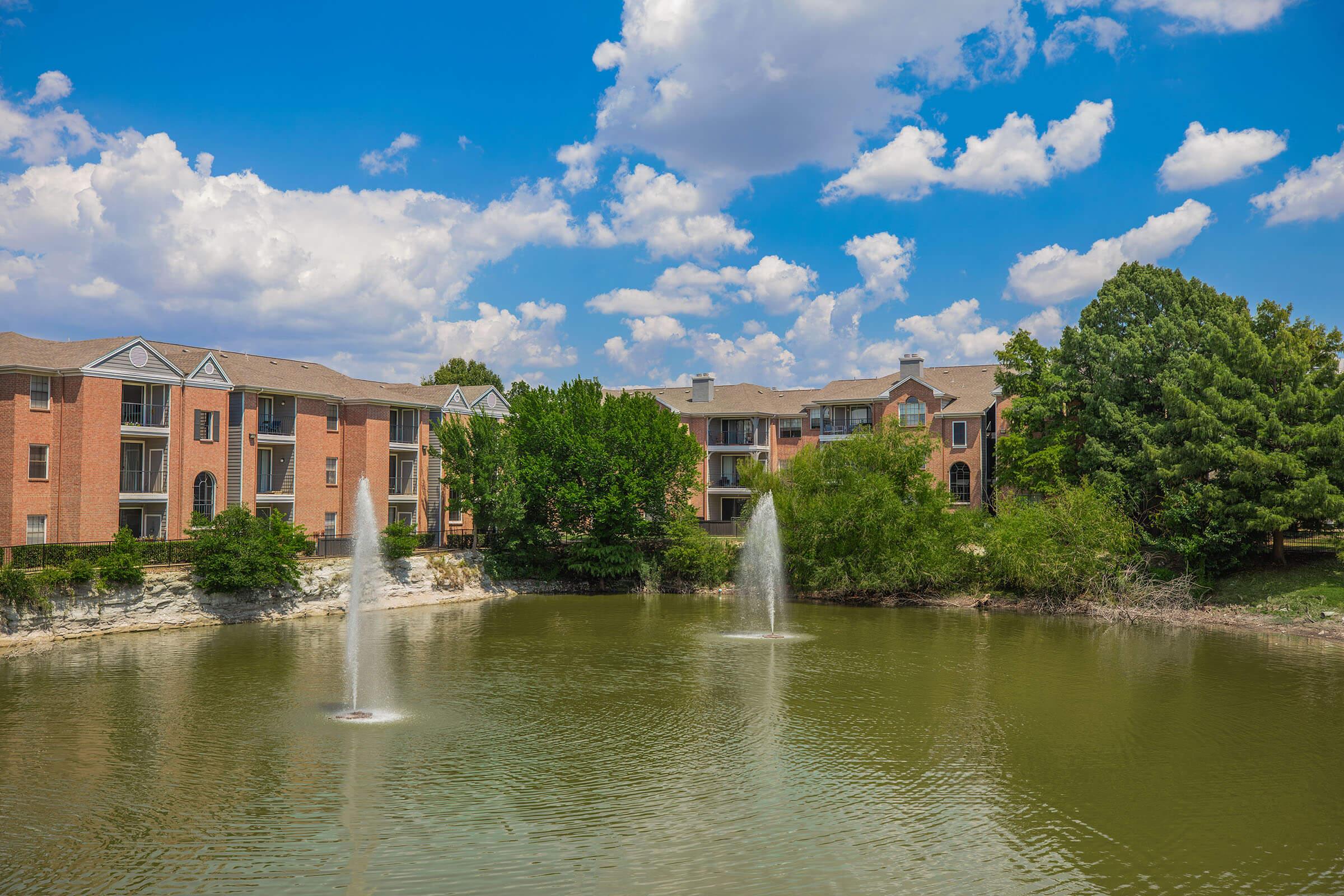 A serene pond in the foreground with two fountains spraying water, surrounded by lush greenery. In the background, red brick apartment buildings are visible under a bright blue sky with fluffy white clouds. The scene conveys a peaceful, residential atmosphere.