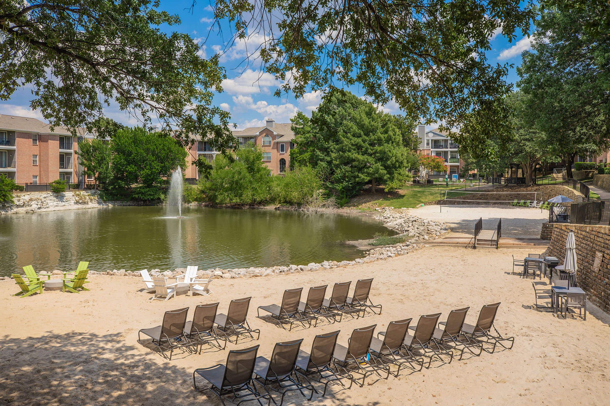 A serene outdoor scene featuring a sandy beach area by a pond. There are lounge chairs arranged in rows facing the water, with a fountain in the pond. Lush greenery and trees surround the area, with residential buildings visible in the background under a clear blue sky.